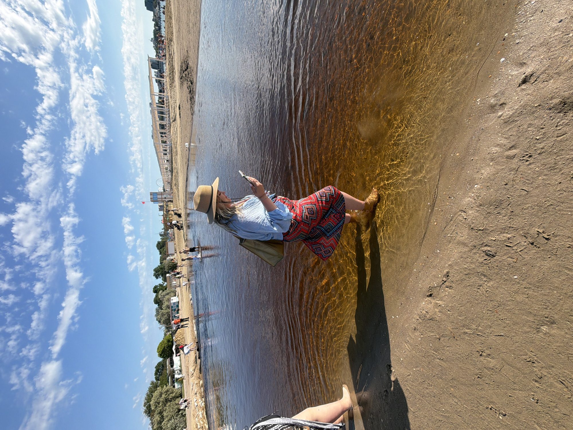 The image, taken in Anapa, Russia, captures a bright, sunny day on a shallow beach or lagoon. The primary subject is a woman standing in clear, shallow water that shimmers with golden sunlight patterns. She wears a light-colored hat, a light blue long-sleeved top, and a vibrant red skirt with an intricate geometric pattern. She is holding a smartphone and appears to be looking at its screen. Her feet are submerged in the water, just barely covered.
To her left, the sandy shore stretches out, meeting the calm water. The sand exhibits fine textures and a few scattered shells or pebbles. In the distance, along the coastline, numerous people are visible on the beach and in the water, enjoying the day. Various beach structures, possibly pergolas or sun shelters, line the shore. Further back, buildings and green foliage indicate a developed coastal area. A Ferris wheel and a red flag can be seen in the background, offering additional location cues.
The sky is a vivid blue with scattered, fluffy white clouds. The overall impression is one of leisure and activity on a warm, pleasant day. There is no visible text in the image.