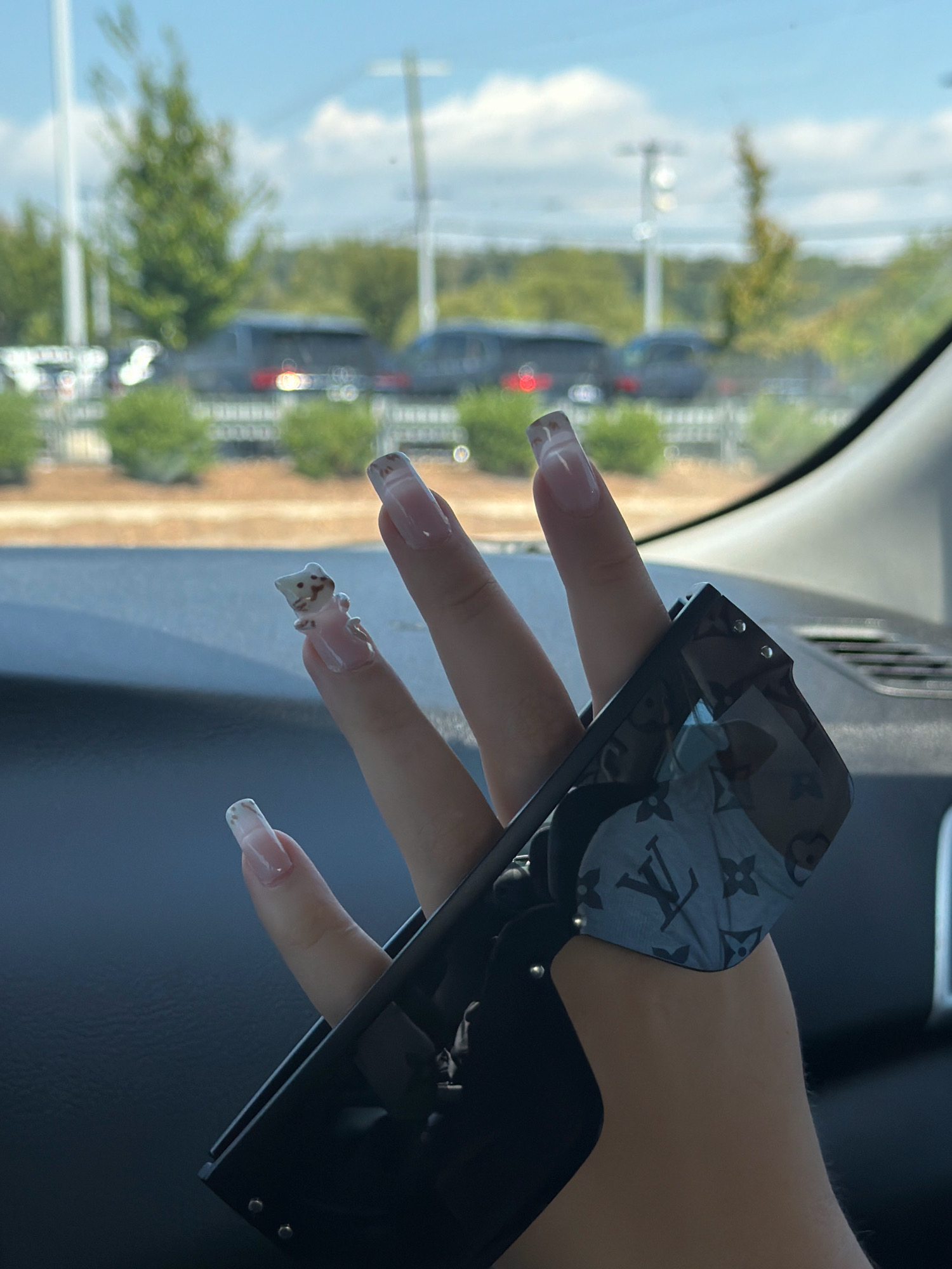 In Asheville, North Carolina, United States of America, a person is shown inside a vehicle, holding a pair of black sunglasses. The sunglasses have a pattern of the Louis Vuitton monogram on the lenses and temples. The person's nails are long, polished, and decorated with a small, three-dimensional charm resembling a cat on the index finger. The background outside the vehicle is blurred, showing trees and a parking lot with several vehicles. The lighting suggests it is daytime.
