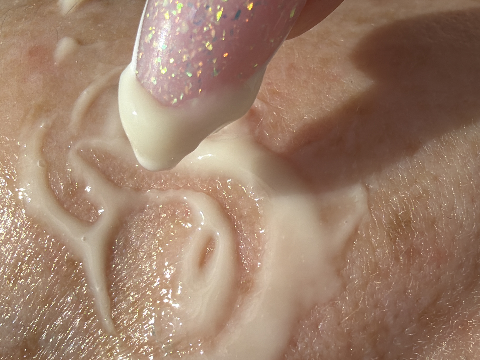A close-up shot shows a light pink fingernail with glitter, coated with a thick white cream or lotion, pressed against human skin. Streaks of the white substance are smeared across the skin's surface, forming swirling patterns. The skin itself appears light-toned and textured, with fine lines and variations in color. A shadow is cast onto the skin, suggesting a light source from above. There is no indication of location or city.