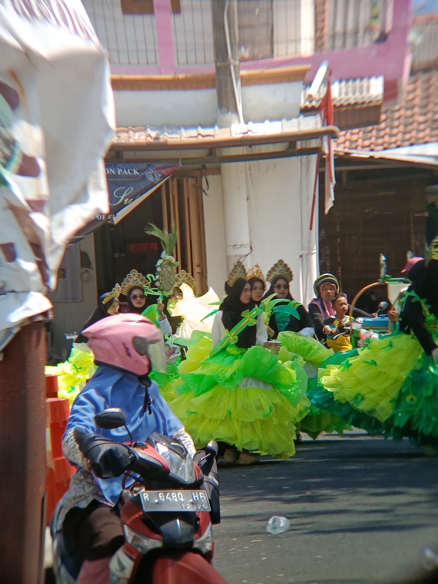 In Desa Kebon Manis, Kecamatan Cilacap Utara, Indonesia, a lively parade is in progress. A person wearing a blue outfit and a pink helmet rides a red and black motorcycle in the foreground, capturing a significant portion of the image. Behind the motorcycle rider, a group of women in traditional attire and elaborate, layered green and yellow dresses are participating in the parade. They are adorned with ornate headpieces, with some wearing hijabs and sunglasses. A young boy in a yellow shirt is also visible amongst the women, riding on a motorcycle. The parade appears to be taking place on a street lined with buildings, suggesting an urban or semi-urban setting. The sunlight indicates it is daytime, casting shadows on the road.