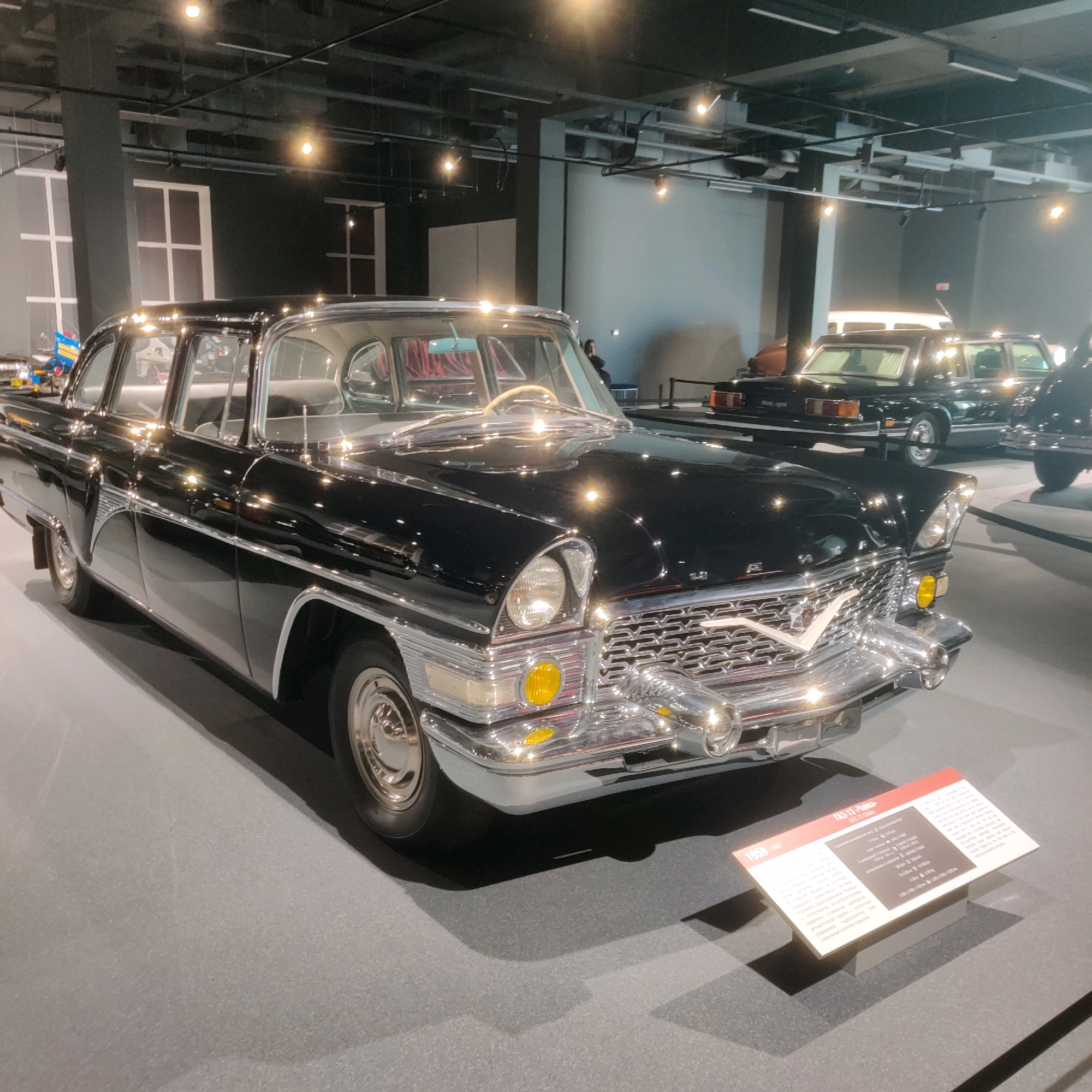 In the city of Verkhnyaya Pyshma, Russia, a black GAZ-13 Chaika sedan is displayed in a museum. The car, manufactured between 1959 and 1982, is positioned at an angle, showcasing its chrome grille, headlights, and side detailing. The interior is visible through the windows, with red velvet upholstery and a cream-colored steering wheel. In the background, other vintage cars, including a black GAZ-11-73 and a limousine, are also on display, illuminated by overhead lighting. A informational plaque with Russian text is placed in front of the GAZ-13. The environment appears to be a modern museum hall with grey floors and dark walls. No people are clearly visible interacting with the vehicles.