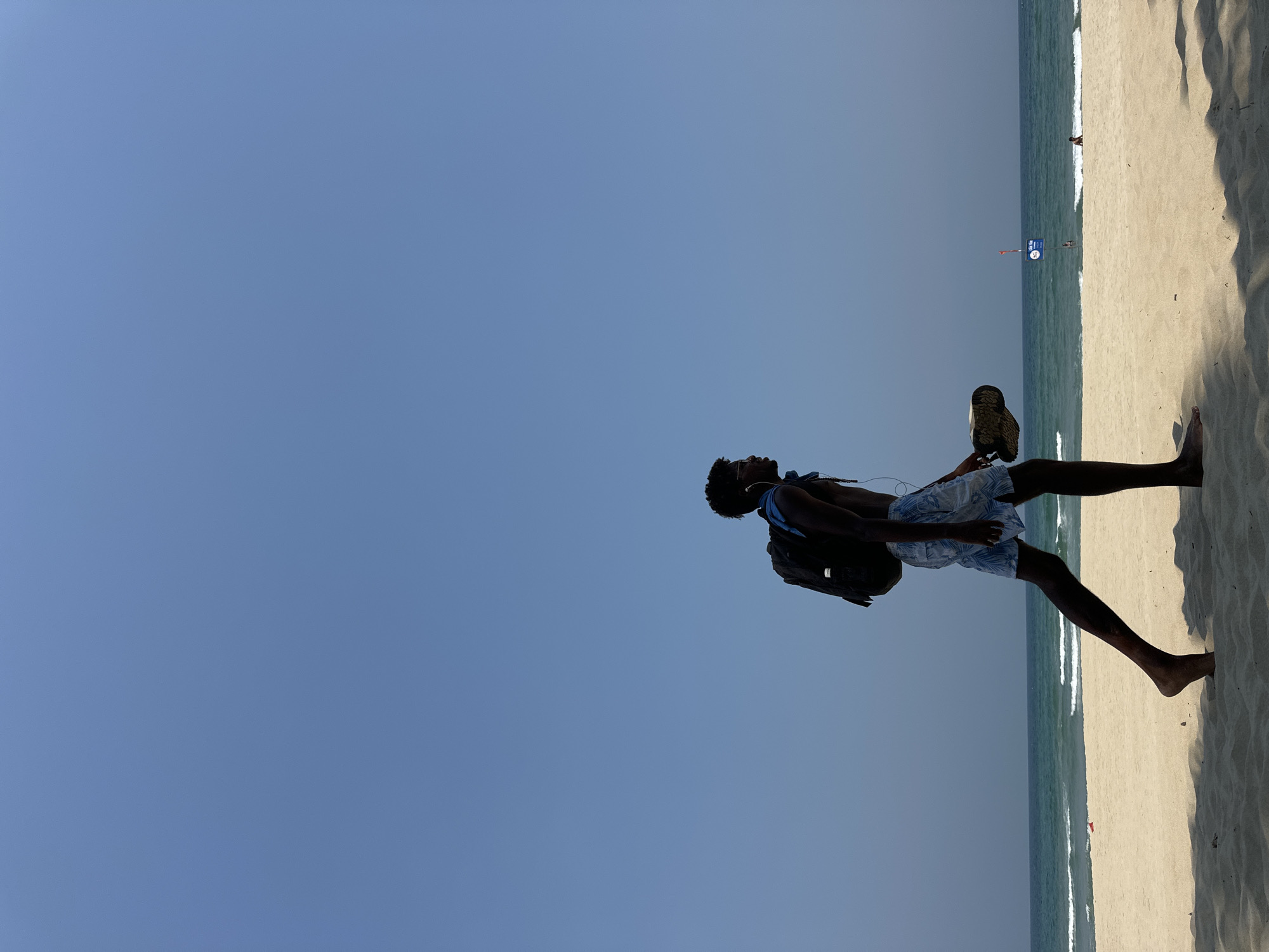 A man stands on a beach. The man is barefoot, wearing shorts, a blue shirt, and a backpack, and is kicking a shoe in the air. Đà Nẵng, Vietnam, is not possible to ascertain from the image.