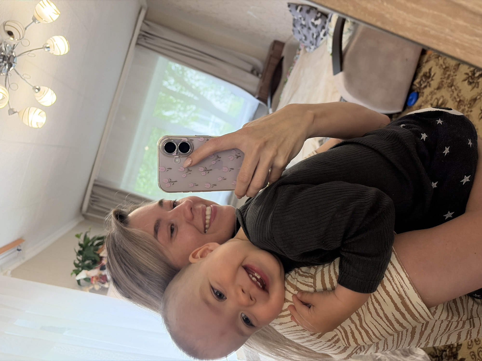 This mirror selfie, taken in a home in Chaikovsky, Russia, captures a joyful moment between a woman and a baby. The woman, with light-colored hair and a warm smile, holds a light purple or pink smartphone with a clear case featuring pink tulip-like flowers, positioned to take the picture. She is wearing a dark, ribbed long-sleeved top.
Cradled in her arms, the baby looks directly at the camera with an exceptionally wide and happy smile, revealing several teeth. The baby also wears a dark, ribbed long-sleeved top, which appears to have white star patterns on the shoulder area, and bottoms with a light brown and white zebra-stripe or abstract pattern. Both subjects radiate happiness and affection.
The setting is an indoor space, likely a living room or bedroom. In the background, a modern chandelier with multiple frosted glass shades is visible. A large window, partially obscured by sheer curtains, shows green foliage outside, indicating it is daytime. Other background elements include wooden furniture, a light-colored chair, and a patterned rug on the floor. The overall impression is one of a loving, cheerful family moment in a bright home environment. There is no visible text in the image.