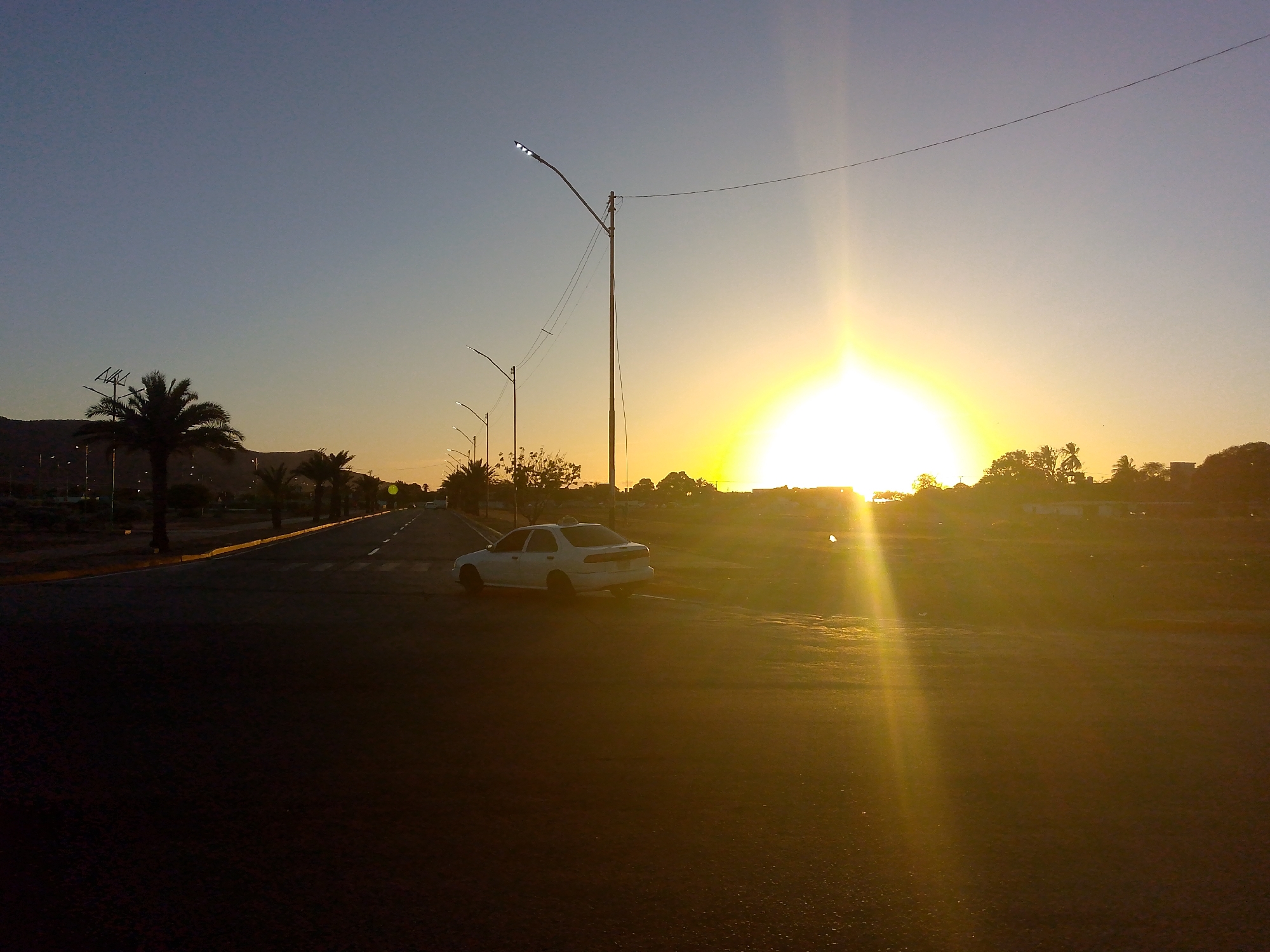 The image shows a street scene in Juan Griego, Venezuela, during sunrise. The sun is positioned directly ahead, creating a strong backlight. A white sedan is parked on the shoulder of the road. The road is paved and appears mostly empty. Streetlights line the side of the road, and palm trees are visible along the left side. In the background, there are houses, trees, and a distant mountain range. The sky is a gradient of blue and orange due to the sunrise.