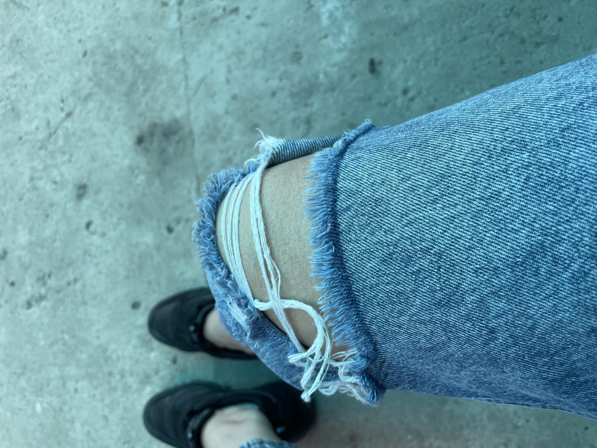 The image depicts a person wearing ripped blue jeans with black shoes standing on a concrete surface. The rips in the jeans expose the skin of the leg and strands of white fabric. The concrete surface has a faded appearance and no specific details indicate the location is Olginka, Russia. No specific actions or interactions are evident.