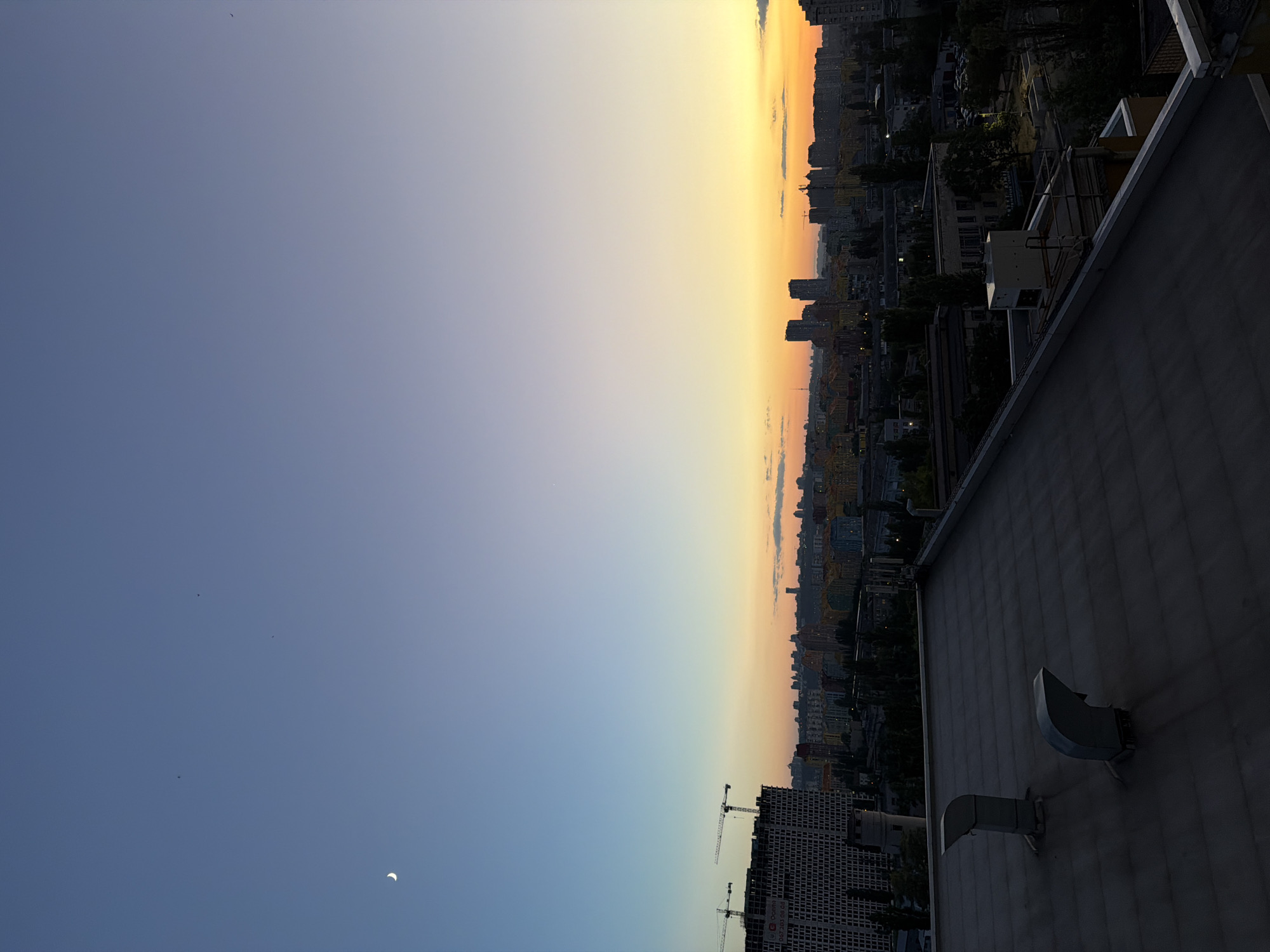 The image depicts a cityscape of Kyiv, Ukraine, during twilight. The sky transitions from a deep blue at the top to a warm orange and yellow gradient near the horizon, indicating either sunrise or sunset. A crescent moon is visible in the upper left quadrant of the sky, along with several small, dark specks that could be birds.
In the foreground, the roof of a building is visible, constructed of dark, weathered material. Two metallic ventilation pipes protrude from the roof, casting shadows.
Further in the distance, the city skyline is dominated by various buildings, including apartment complexes and office structures. Two prominent construction cranes are visible on the left side, atop a large, unfinished building. The buildings display a mix of architectural styles and colors, with some appearing modern and others older. Trees are scattered throughout the cityscape, particularly in the middle ground, their dark foliage contrasting with the illuminated sky. The overall impression is an urban environment at the cusp of day and night.