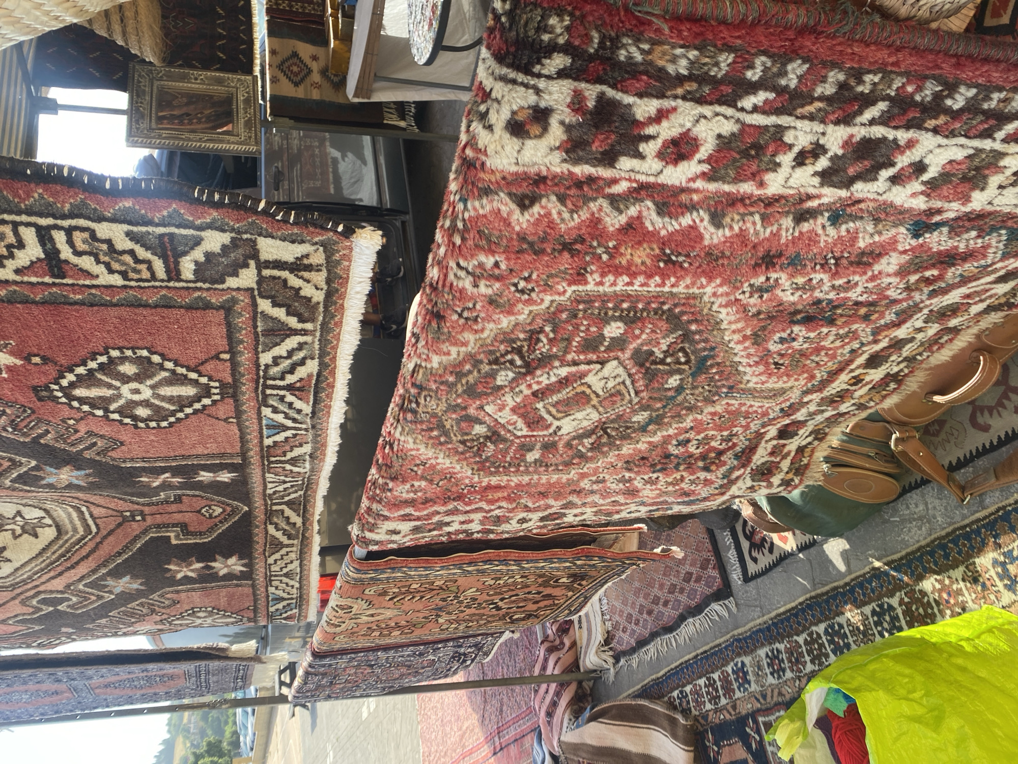 An outdoor market stall in Manilva, Spain, displays a variety of intricate, handmade rugs. The rugs are primarily red, brown, and cream, featuring traditional geometric patterns and floral motifs. Some rugs are hung from a metal rack, while others are laid out on the ground. In the background, a vehicle with an open door is visible, along with a framed mirror leaning against it. A table with a colorful, mosaic-patterned top sits to the right, suggesting a seating area. A bright green bag filled with yarn and a few leather bags and a stool are also present, adding to the market atmosphere. The setting appears to be paved, with a glimpse of a hillside and trees in the distance.