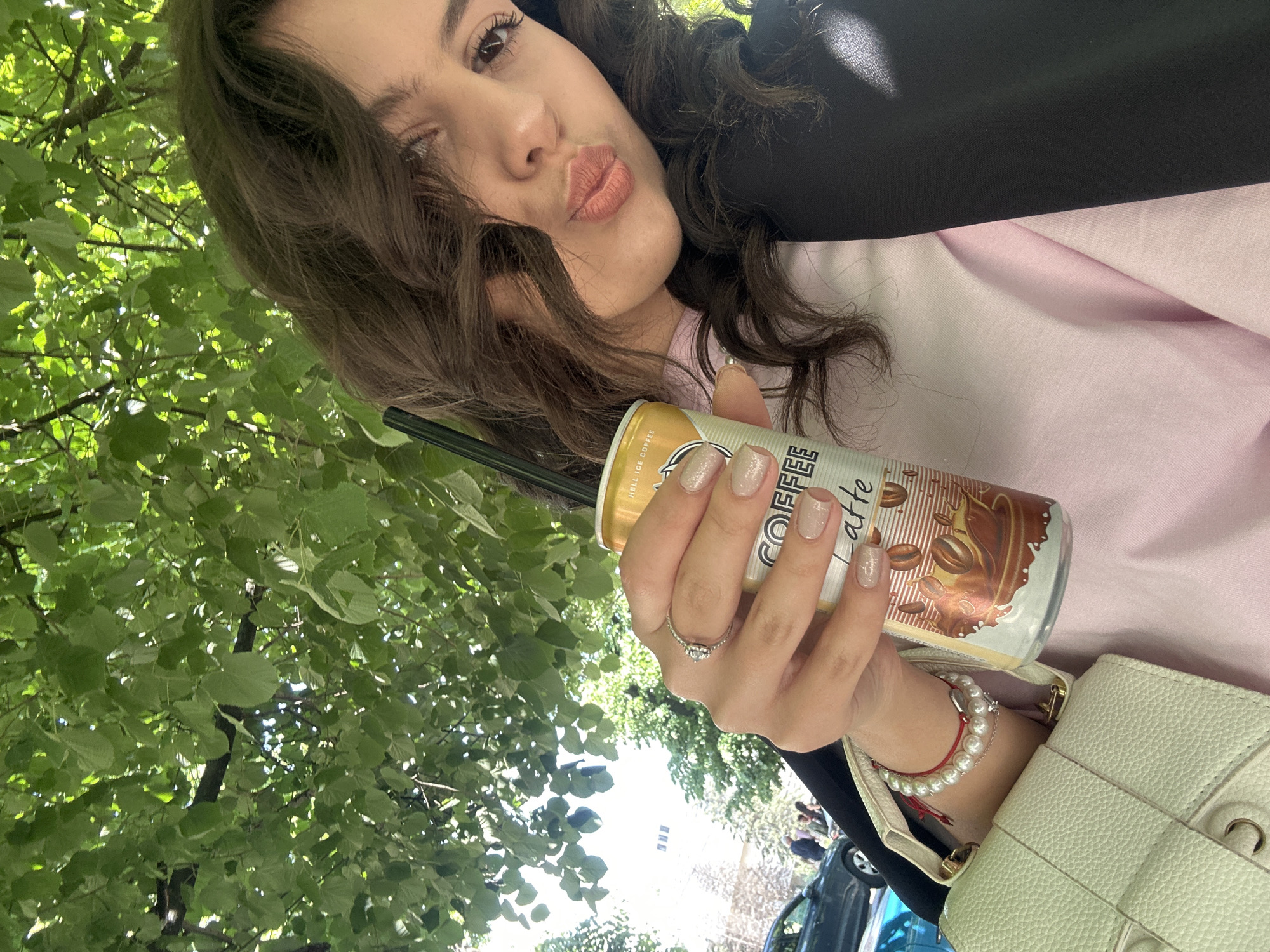 A young woman takes a selfie while holding a can of iced coffee. She has dark hair and is wearing a pink shirt, a black jacket, a ring, and a bracelet. The can is labeled "COFFEE latte" and has a brown and gold design. A straw is inserted into the can. The background includes green leaves from a tree and a glimpse of a car and a building. The location is an outdoor area in a city; Unknown, Unknown.