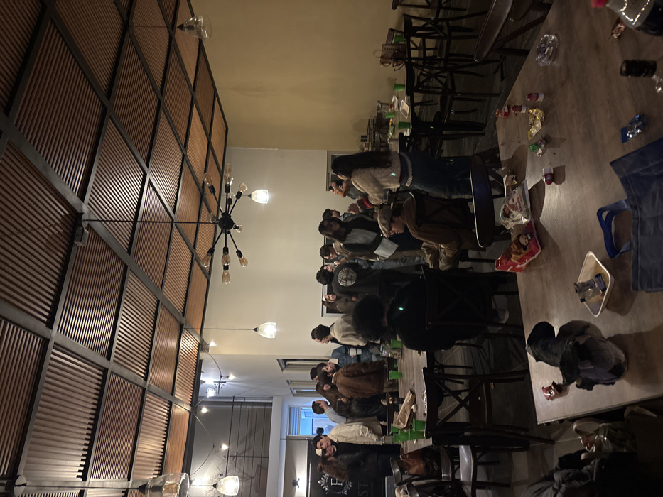 A long table occupies a room, littered with food wrappers and discarded items. People sit along the table, engrossed in an activity. One side of the room features a wall covered in a grid pattern of wooden slats. Chandeliers hang from the ceiling, providing light. Chairs surround the table, and additional furniture and belongings are visible near the edges of the image. The overall atmosphere suggests a casual gathering in a slightly cluttered space.