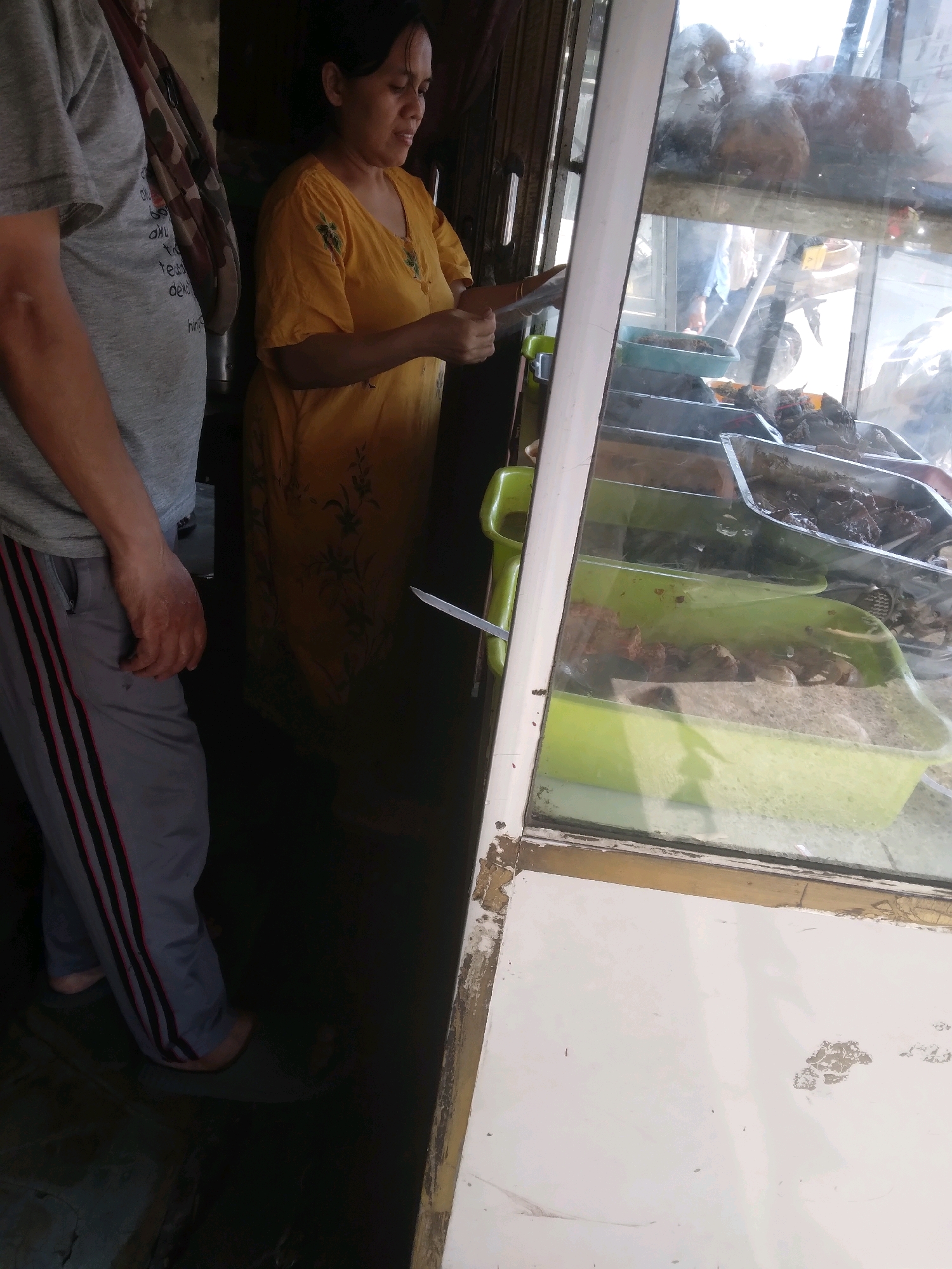 A woman in a yellow dress stands behind a display case filled with food. To her left, a person in a grey t-shirt and grey sweatpants with red and black stripes stands with their back partially to the camera. The display case has glass windows and contains several trays of food, including what appears to be meat and poultry. A green plastic handle protrudes from the side of the display case. The scene is indoors, likely a food stall or market. The lighting is dim, suggesting it may be inside a building or covered area. The image appears to be taken in Purwokerto, Indonesia, as indicated by the context.