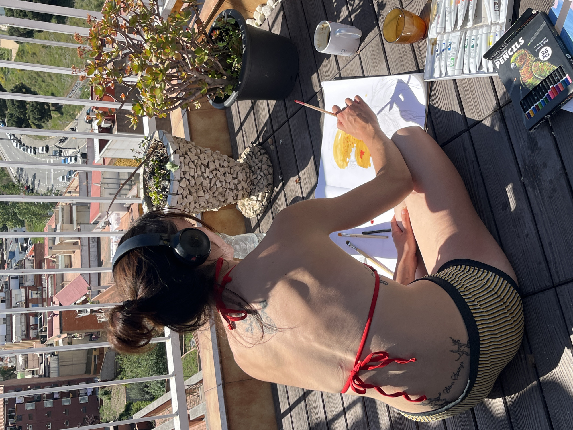 A woman wearing headphones, a red bikini top, and striped bikini bottoms is seated on a wooden deck and painting in a sketchbook. She is surrounded by art supplies, including a set of coloring pencils, tubes of paint, and paintbrushes. Nearby are two potted plants, a white mug, and a glass. The scene is outdoors, likely on a balcony, with a cityscape visible in the background. Based on the architecture and layout of the buildings, the location appears to be in the city of Barcelona, Spain.