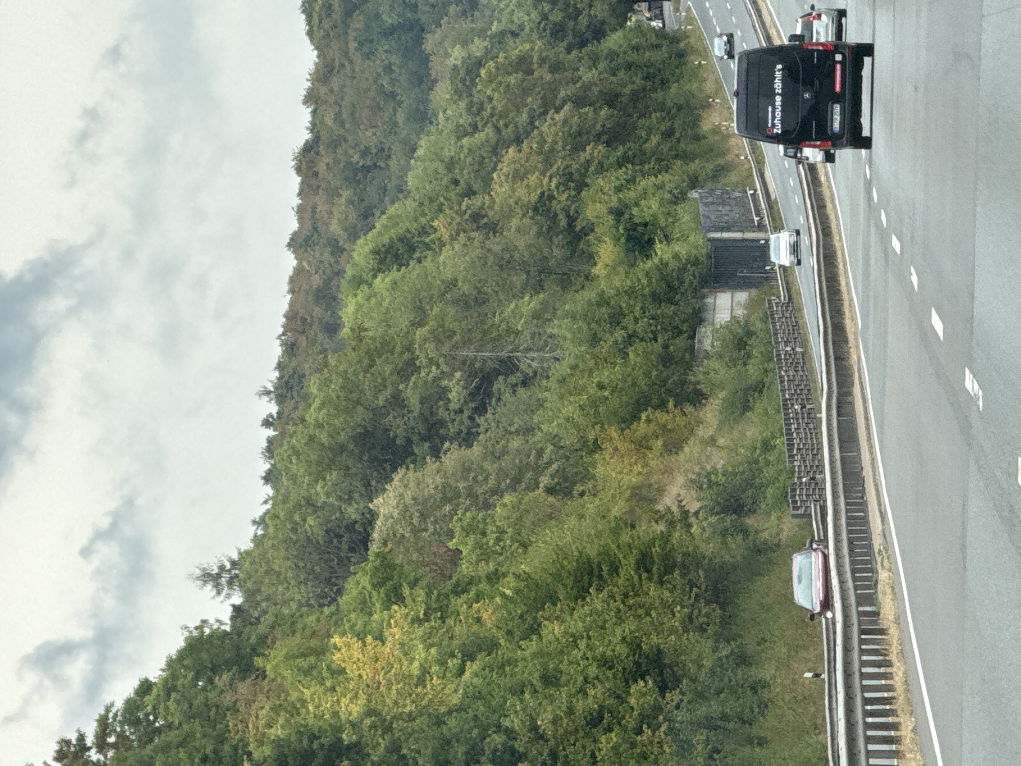 The image depicts a view of a highway in Herzberg, Germany, on a cloudy day. The highway has multiple lanes and a barrier separating the lanes of traffic from the adjacent wooded hillside. Several vehicles are visible on the highway, including a black van with the text "Zuhause zählt's" on its rear, a red car stopped on the shoulder, and other cars in motion. The wooded hillside is covered in a variety of trees, some of which show signs of autumn color. The sky is overcast with gray clouds. The overall scene conveys a sense of travel on a German highway, surrounded by natural landscape.