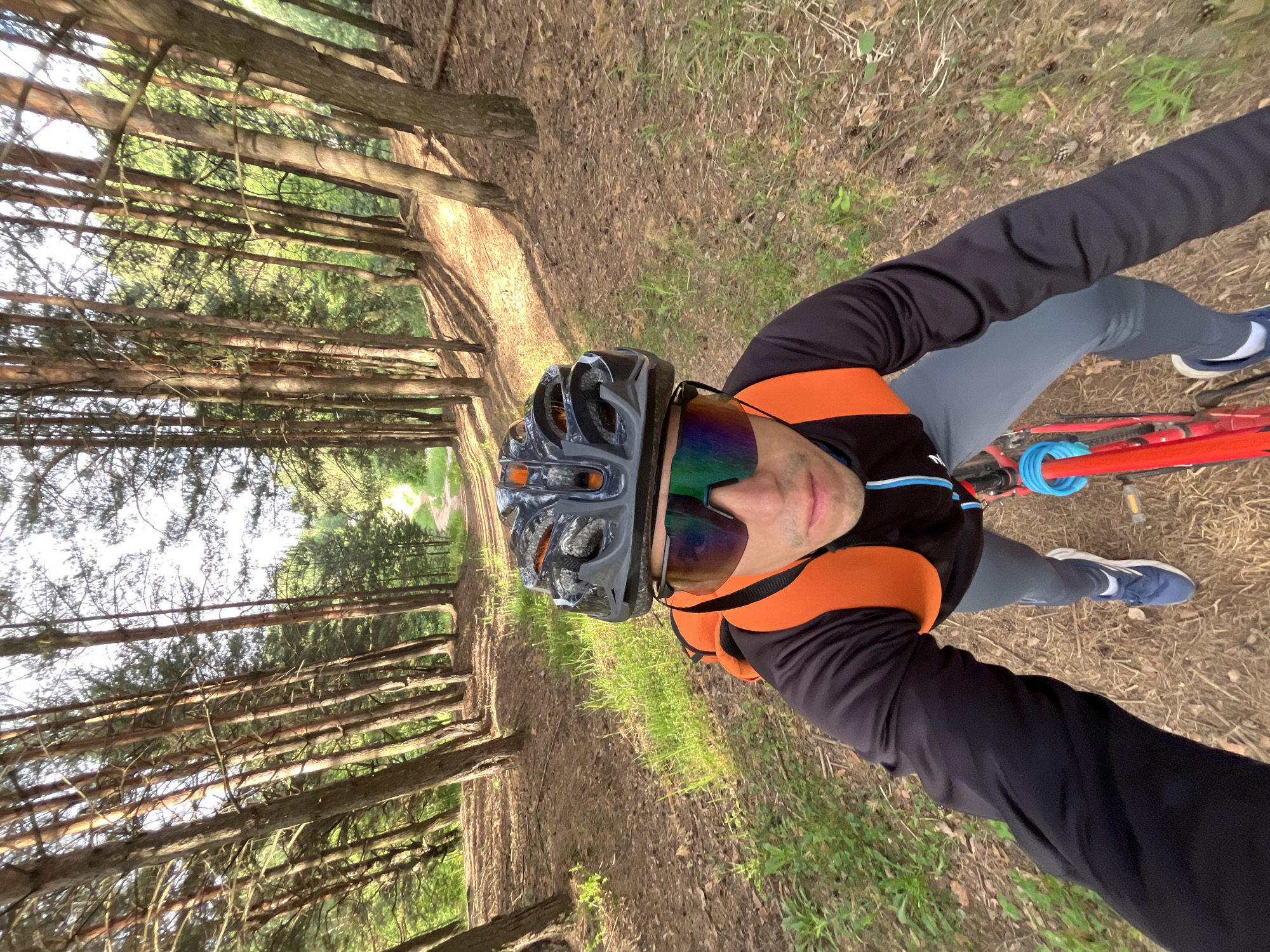 The image shows a first-person perspective of a person riding a bicycle on a dirt path through a pine forest. The person is wearing a black helmet with orange accents, large reflective sunglasses with a green and purple tint, a black long-sleeved shirt, and grey leggings. They have an orange backpack with black straps. The bicycle is red. The path is narrow and surrounded by tall pine trees with sunlight filtering through the canopy, creating dappled shadows on the ground. The ground is covered with pine needles and some patches of grass. The overall environment suggests a rural or natural setting, likely on the outskirts of Chaikovsky, Russia, given the characteristics of the pine forest and the visible vegetation. The person appears to be actively engaged in cycling.