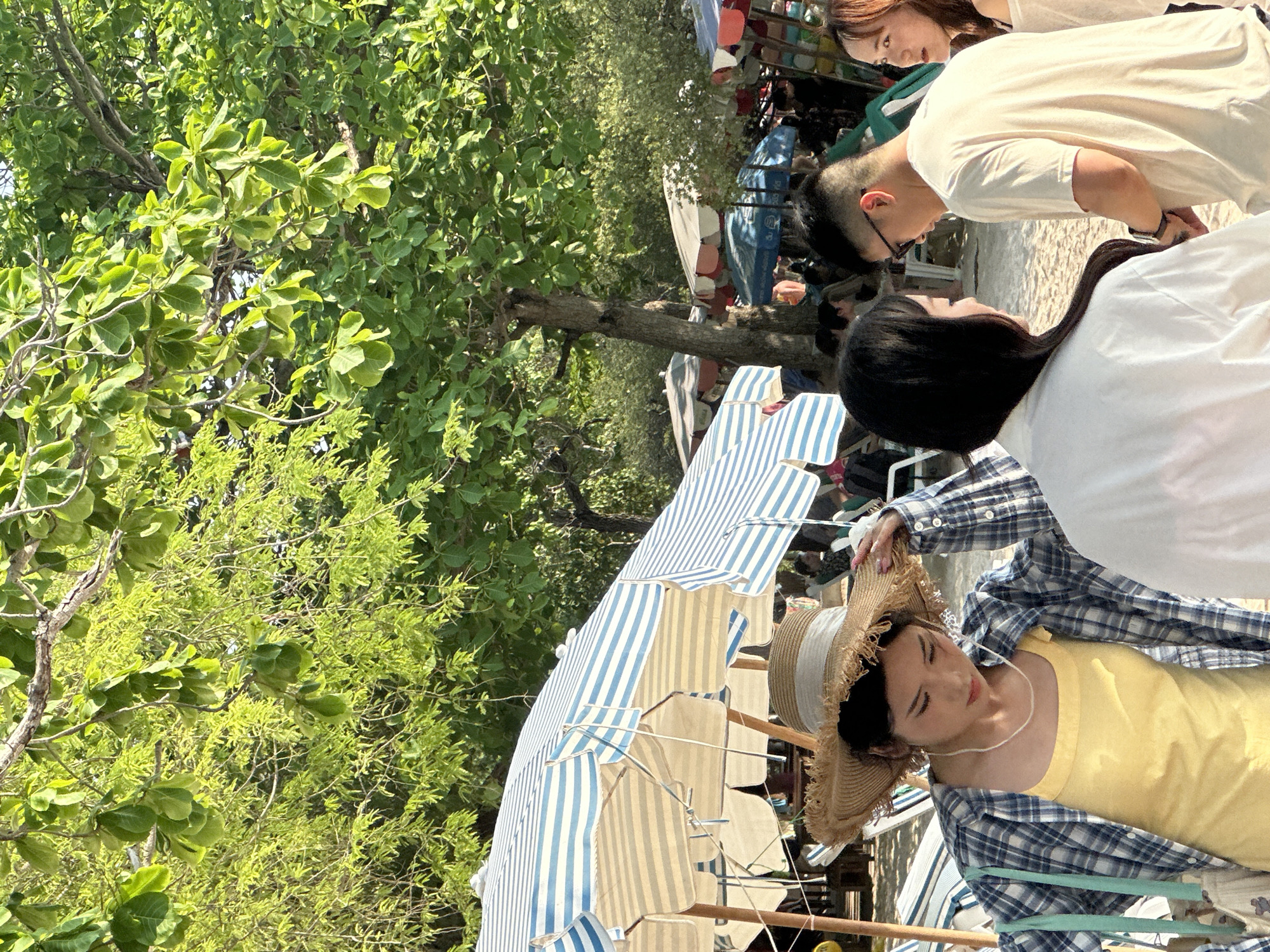 The image captures a scene on a sunny day in Pattaya City, Thailand. The focus is on a young woman wearing a yellow top, a blue and white checkered shirt, and a straw hat. She is seated under a blue and white striped beach umbrella. Nearby, there are three people standing and interacting with each other. Lush green trees are visible in the background. The location appears to be a beachside setting with numerous umbrellas, indicating a public area.
