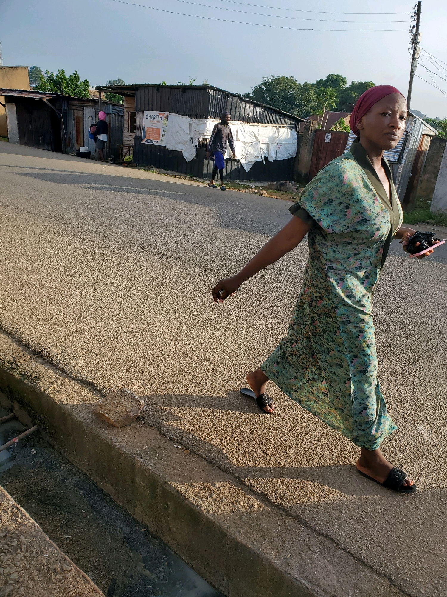 A medium shot shows a woman walking on a dusty road in the city of Jos, Nigeria. She is wearing a green floral dress and a maroon headscarf. Her body is angled towards the viewer, with her right arm extended slightly and her left arm holding a pink smartphone and a black object. She is wearing black sandals with a logo. In the background, several corrugated iron structures are visible, some with white tarpaulin coverings. A sign with "CHERITH Kitchen & Catering" is attached to one of the buildings. Another person in a dark shirt and blue shorts is walking away from the camera in the distance. The road surface is asphalt with visible texture and debris. A concrete drainage ditch runs along the side of the road, containing murky water and debris. The sky is clear and pale blue. The sun is casting long shadows.
