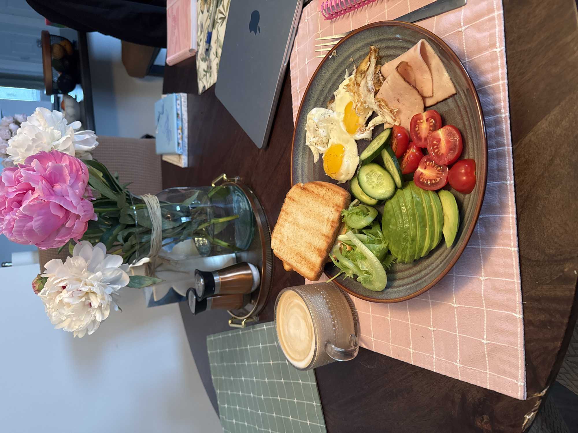 A top-down, slightly angled shot shows a breakfast spread on a dark wooden table, likely in a home dining area. The meal is presented on a grey, speckled plate and consists of two fried eggs, sliced ham, halved cherry tomatoes, cucumber slices, sliced avocado, and a small pile of mixed greens. To the side of the plate is a piece of toasted bread. A glass mug filled with coffee and latte art sits to the lower right of the plate, on a green checkered placemat. A pink checkered placemat lies beneath the plate and coffee cup.
To the left of the table setting, a bouquet of pink and white peonies is arranged in a clear glass vase. A brass tray with two small spice shakers is behind the vase. In the background, a glimpse of a laptop with an Apple logo is visible, along with several small notebooks. The overall impression is that of a leisurely breakfast or brunch. The lighting suggests it could be daytime, possibly morning or early afternoon. There are no people visible in the image.