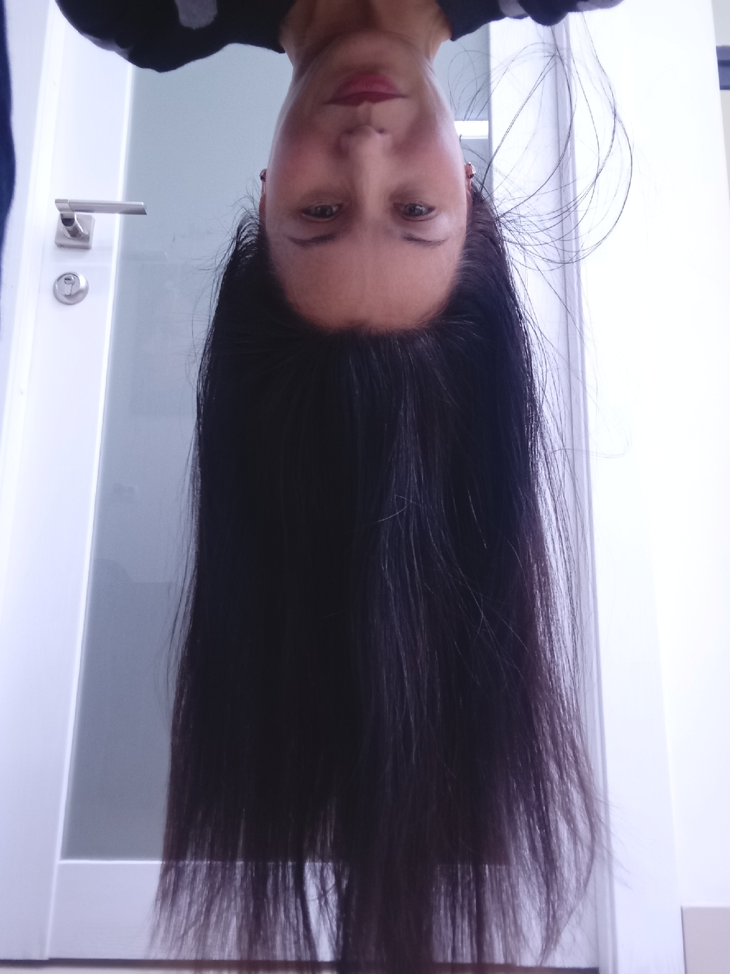 This full-shot, inverted image features a woman looking downwards, appearing upside down to the viewer, with her long, straight, dark hair flowing freely towards the bottom of the frame due to gravity. Her face shows a neutral expression with slightly parted lips highlighted with a reddish hue. Her eyes are open, and a small earring is visible on her right ear. She wears a dark garment with some lighter markings visible on her shoulder and upper arm.
The background primarily consists of a white interior door frame on the left side, featuring a modern, silver-toned horizontal door handle and a matching keyhole or lock mechanism below it. Behind the door frame, a translucent or frosted glass panel is visible, suggesting a well-lit space beyond. The lighting throughout the image is bright and even, indicating either ample daylight or strong indoor illumination. The woman's hair extends almost the entire length of the lower image. There is no visible text. The image's location context is Izhevsk, Russia.