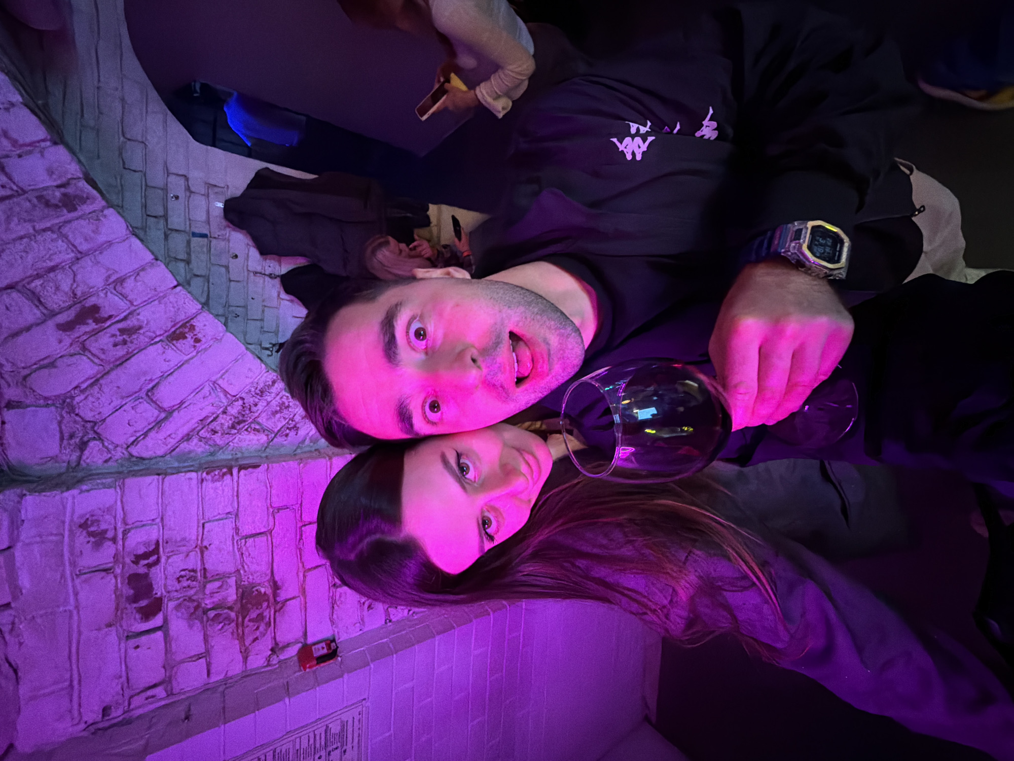 The image captures two individuals, a man and a woman, posing in an indoor setting bathed in vibrant purple and magenta lighting, suggestive of a bar, club, or lounge. The photo is taken from a high angle, with the subjects appearing to be lying down or leaning back against a surface, looking up directly at the camera.
In the foreground, a man with dark hair and stubble displays a look of surprise or playful excitement, with wide eyes and an open mouth. He wears a black long-sleeved shirt featuring the distinctive white Kappa logo on his right arm. A prominent digital wristwatch, resembling a G-Shock with a clear or metallic bezel, is visible on his left wrist. His left hand holds a wine glass.
Positioned below and beside him, a woman with long dark hair rests her head, looking towards the camera with a subtle, gentle smile that conveys contentment.
The background shows a white brick wall, parts of which reveal underlying red brick, all illuminated by the intense purple light. Below the brick wall, a white tiled surface, possibly a bar counter or lower wall, is visible. On this surface, a small red object, possibly an electrical component or alarm, and a partially obscured document with unreadable text can be discerned. Further in the background, faint blue light and blurry figures suggest other people and elements of the venue.
The overall atmosphere is lively and intimate, characteristic of a nightlife environment. The dramatic colored lighting dominates the scene, highlighting the subjects' expressions and setting a clear nighttime mood for a social gathering. The man's shirt features a visible Kappa logo.