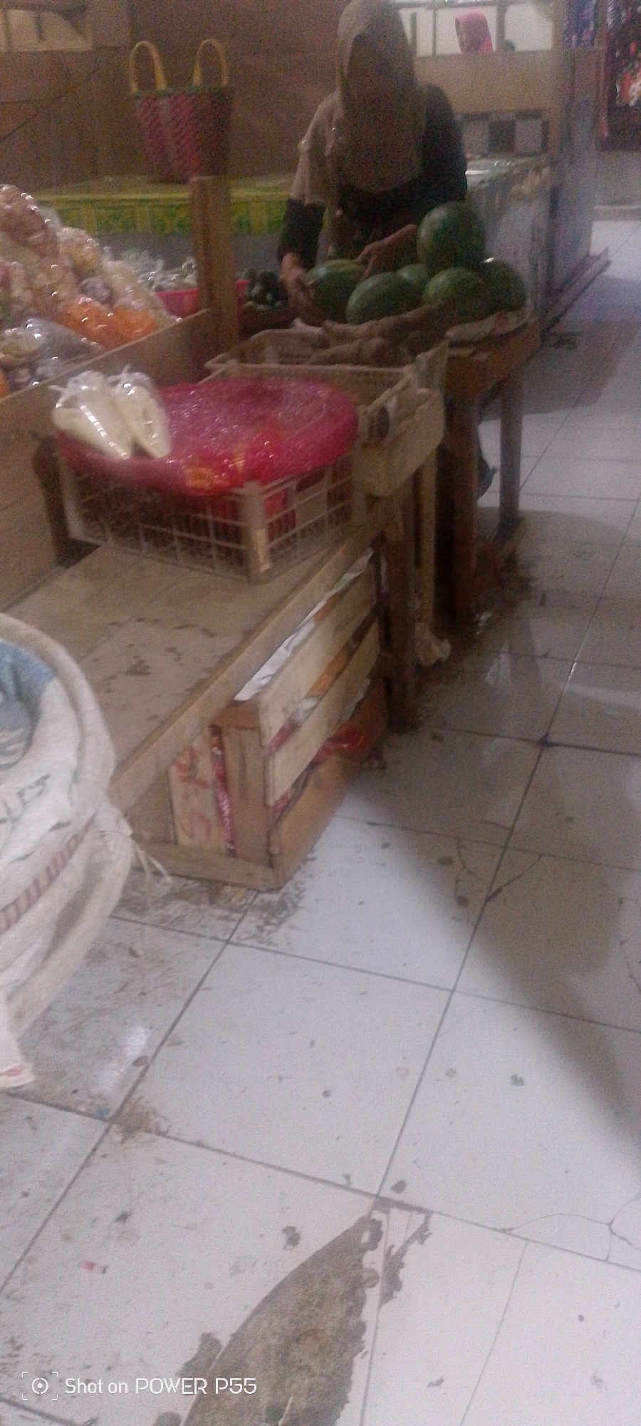 An overhead shot shows a person wearing a headscarf and apron standing behind a market stall. They are sorting through what appears to be fruit, including large green watermelons. The stall is made of wood and has several crates filled with produce. In the foreground, a large white sack lies on the floor. The floor itself is tiled and appears somewhat dirty, with some debris scattered around. The overall lighting is dim, suggesting an indoor market. The location is identified as Unknown, Unknown.