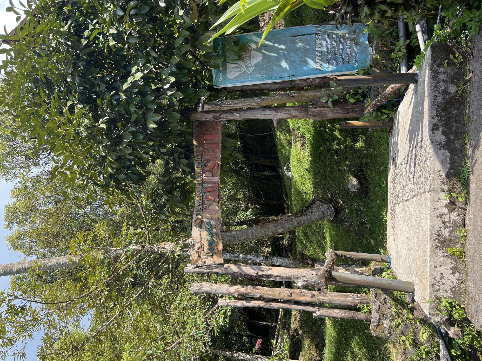 The image shows an entrance to a campsite in Sukasada, Indonesia. A rustic wooden sign with the words "JEMBONG CAMPSITE" is mounted on wooden posts. To the right of the wooden sign, a blue tarp sign lists activities offered at the campsite. The entrance is made of concrete, leading into a grassy area surrounded by lush green trees and foliage. The overall scene depicts a natural, outdoor environment.