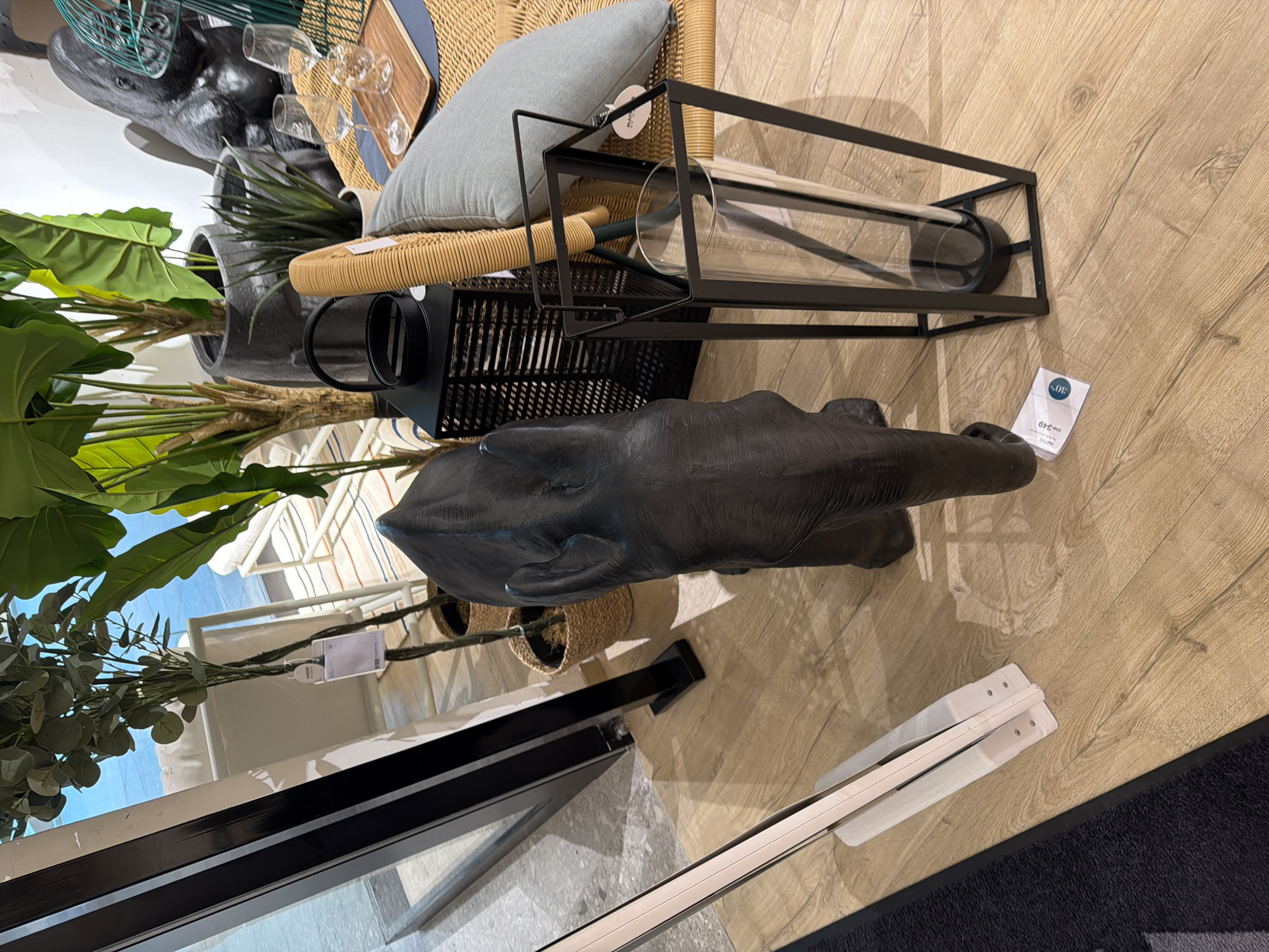In an indoor retail environment, a dark, textured elephant statue stands on a light-colored wooden floor. To the right of the elephant is a tall, black metal lantern with a glass cylinder. In the background, there are various decorative plants in woven baskets, a black gorilla statue, and a table with glassware and a wooden tray. A black frame with a glass panel runs along the left side of the image, suggesting a window or display case. A white, folded object with a dark accent is positioned on the floor in the lower left quadrant.