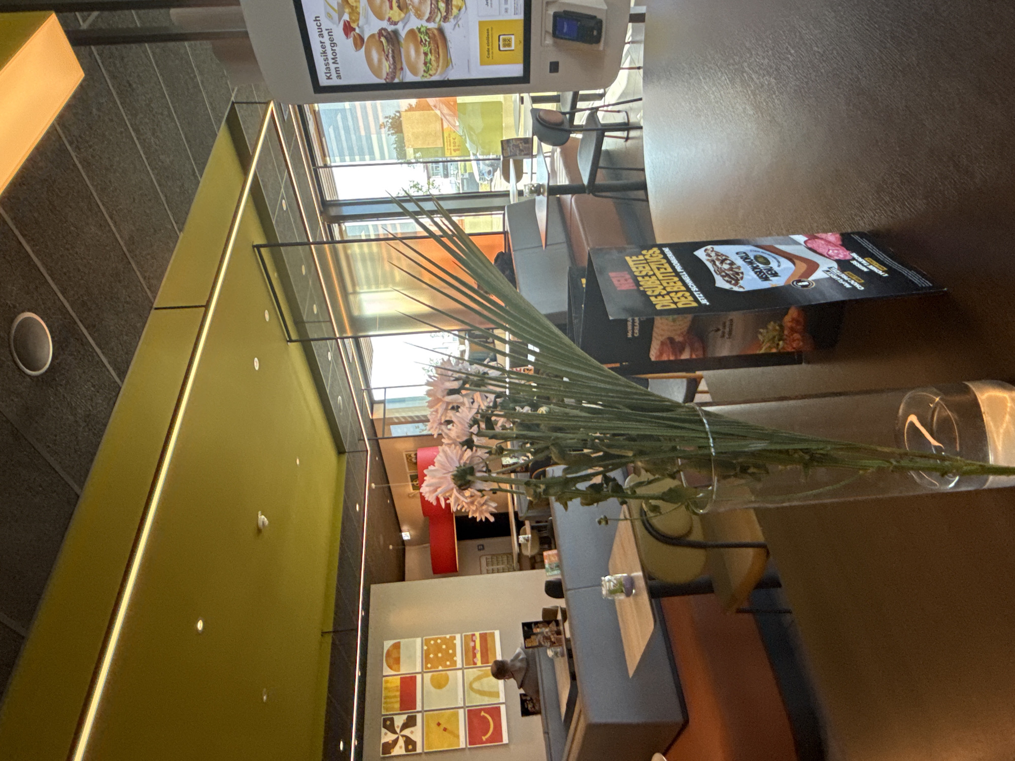The image captures the interior of a McDonald's restaurant in Werther, Germany, with a focus on a table in the foreground adorned with a vase of light pink flowers and a menu. The ceiling is a striking yellow with integrated strip lighting and darker acoustic panels. In the background, a person is seated at a booth, facing a wall decorated with a grid of nine colorful square artworks. Large windows offer a view of the exterior, suggesting a bright, sunny day. A digital menu board displays images of burgers and text in German. The overall atmosphere is modern and clean, with a mix of seating arrangements including booths and individual tables.