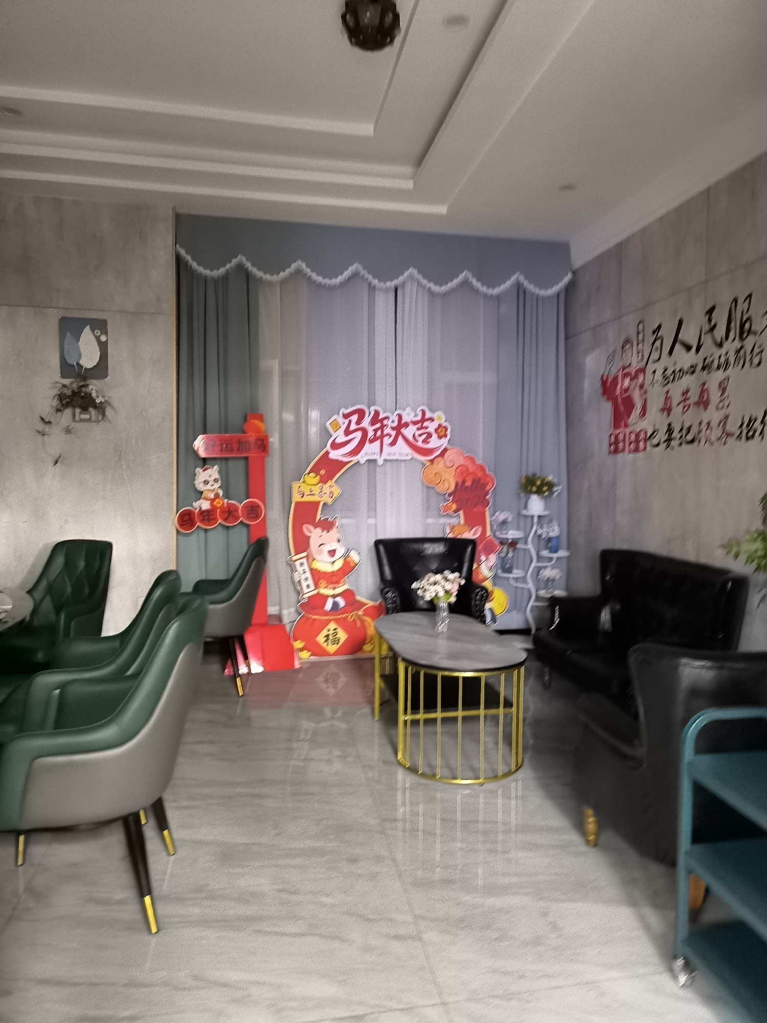 The image depicts an indoor scene that appears to be a waiting area or a small lounge, decorated for the Chinese New Year. In the center of the room, there is a large, arch-shaped decoration with a cartoon pig and auspicious Chinese characters, possibly signifying good fortune. To the left, a red signpost with a pig decoration stands near several green chairs. Further to the left, a framed picture of leaves is mounted on a grey wall. The background features sheer white curtains and a light blue valance.
In the foreground, a modern coffee table with a grey top and a gold metal base is positioned in front of a black leather sofa. A vase with flowers sits on the coffee table. To the right of the sofa, a dark green multi-tiered shelving unit is partially visible. The flooring is a light-colored, marble-patterned tile. On the far right wall, there is a mural with red and black Chinese calligraphy and illustrations.
While the image is of an indoor setting, it is difficult to ascertain the specific location in Lucaogou Town, China, without further context or external information. The scene is static, with no visible people or actions taking place. The overall impression is one of a festive and modern interior designed for customer comfort and seasonal celebration.