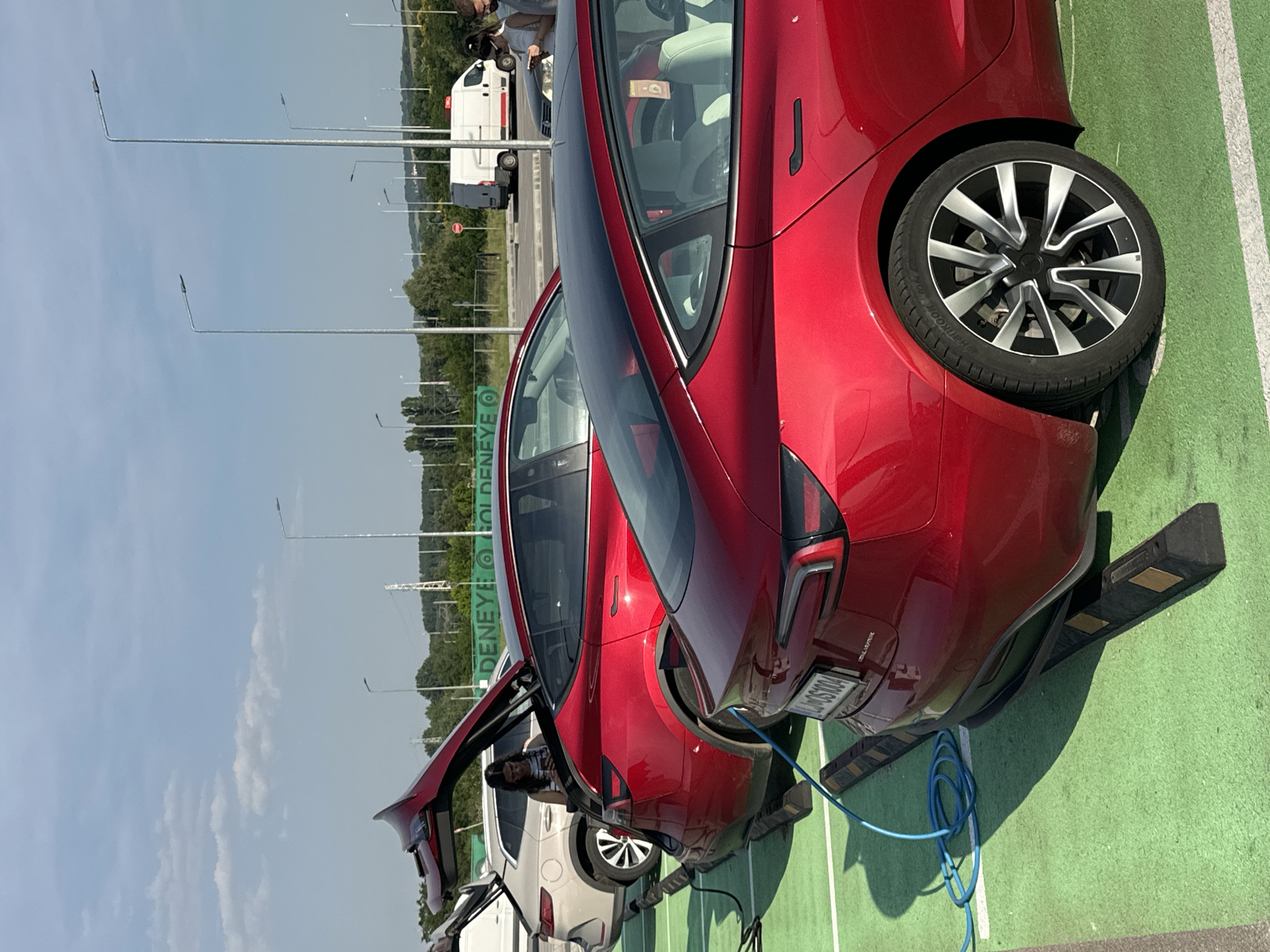 This detailed description outlines a scene at an electric vehicle charging station located at the Kapitan Andreevo border crossing in Bulgaria, evidenced by the "DENEYE GOLDENEYE" signage visible in the background.
The image is captured outdoors under a clear blue sky with scattered cumulus clouds, illuminated by direct sunlight. The ground surface consists of a vibrant green material, likely painted asphalt or artificial turf, marked with white lines defining individual parking bays. Each bay is equipped with black and yellow striped parking stoppers.
Three vehicles are prominent in the midground and foreground. The central vehicle is a red Tesla Model 3, parked facing away from the viewer with its trunk open. A coiled blue electric charging cable is connected to its rear-left charging port, extending towards the left side of the frame where the charging apparatus is partially obscured. The license plate "L4-OS1004" is visible on this Tesla. A woman with dark hair and glasses, wearing a striped top, stands near the open trunk, looking down at a mobile phone in her hands. Directly behind this Tesla, a light beige SUV with a multi-spoke wheel design is also parked with its trunk open.
In the foreground, on the right side of the image, the rear section of another red Tesla Model 3 is visible. This vehicle displays a rear wheel with black and silver multi-spoke rims and features light-colored interior seats, visible through the rear window.
The background reveals a landscape of rolling hills covered in trees. Numerous tall, slender streetlights are interspersed across the area, alongside visible power lines and utility poles. A large green sign prominently displays "DENEYE" and "GOLDENEYE" in white lettering. On a road beyond the charging station, a white delivery van with red rear markings is visible. A red circular stop sign is partially visible near the road. On the far right edge of the frame, two individuals are partially visible, appearing to be standing near a dark vehicle. One person, wearing a white top and a face mask, faces left towards the scene.