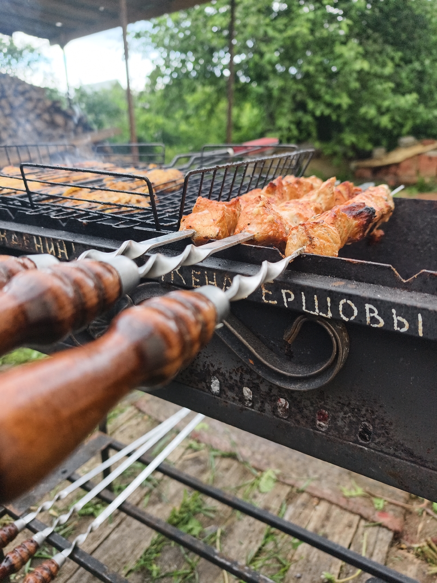 The image depicts an outdoor grilling scene in Prikamskii, Russia. In the immediate foreground, two metal skewers with dark, worn wooden handles are held over a dark metal brazier. Each skewer has a spiraled metal shaft and holds multiple pieces of cooked meat, likely shashlik, showing browning and some charring. Behind the skewers, a rectangular metal grill rack within the brazier holds numerous smaller pieces of cooked, golden-brown food, possibly chicken or fish, from which light smoke is visibly rising.
The brazier itself is made of dark metal and features white stenciled or painted Russian text. The left side of the brazier shows the partial inscription "КЛЕНИН" (KLENIN), while the right side clearly displays "ЕРШОВЫ" (YERSHOVY). The background is an out-of-focus outdoor environment dominated by lush green trees and foliage under a bright, possibly overcast, sky. A light-colored structure, possibly a canopy or pergola, is visible overhead. The ground beneath the brazier consists of weathered wooden planks interspersed with sparse green vegetation. No individuals are present in the frame.