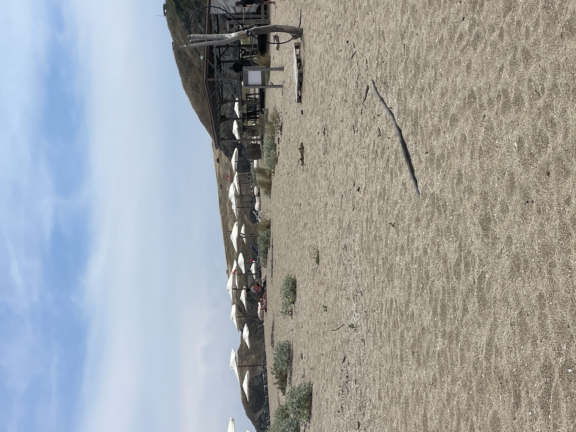 The image depicts a beach scene in Sinemorets, Bulgaria. In the foreground, a sandy beach is visible with scattered driftwood and small vegetation. A row of white umbrellas and beach chairs are set up further back, suggesting a designated beach area. To the right, a wooden structure, possibly a beach bar or restaurant, is situated against a grassy hillside. Several people are present, some lounging under umbrellas and others walking or standing near the bar. The sky is partly cloudy, with a blue expanse above. The overall impression is that of a serene, sunny day at a seaside resort.