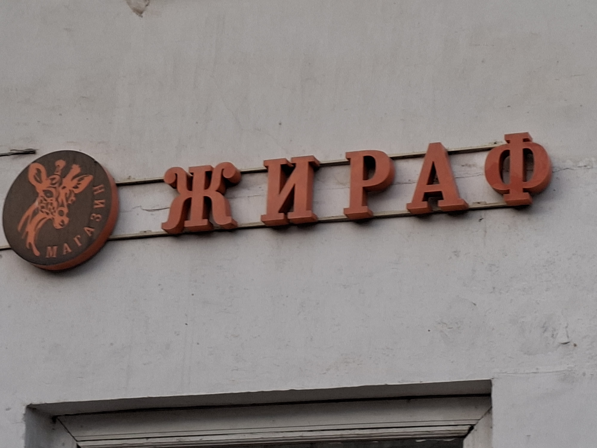 A close-up, eye-level, exterior shot shows an orange sign with a stylized giraffe head and the Russian word "МАГАЗИН" (shop) in Cyrillic script on a circular brown background to the left. To the right of the giraffe sign, in orange 3D Cyrillic letters, is the word "ЖИРАФ" (giraffe), which appears to be the name of the shop. The signs are mounted on a light-colored, rough-textured wall, likely concrete or stucco, above a darker horizontal line, possibly part of a window frame or architectural element. The background is out of focus. The lighting suggests it is daytime, but overcast. The scene is devoid of people.