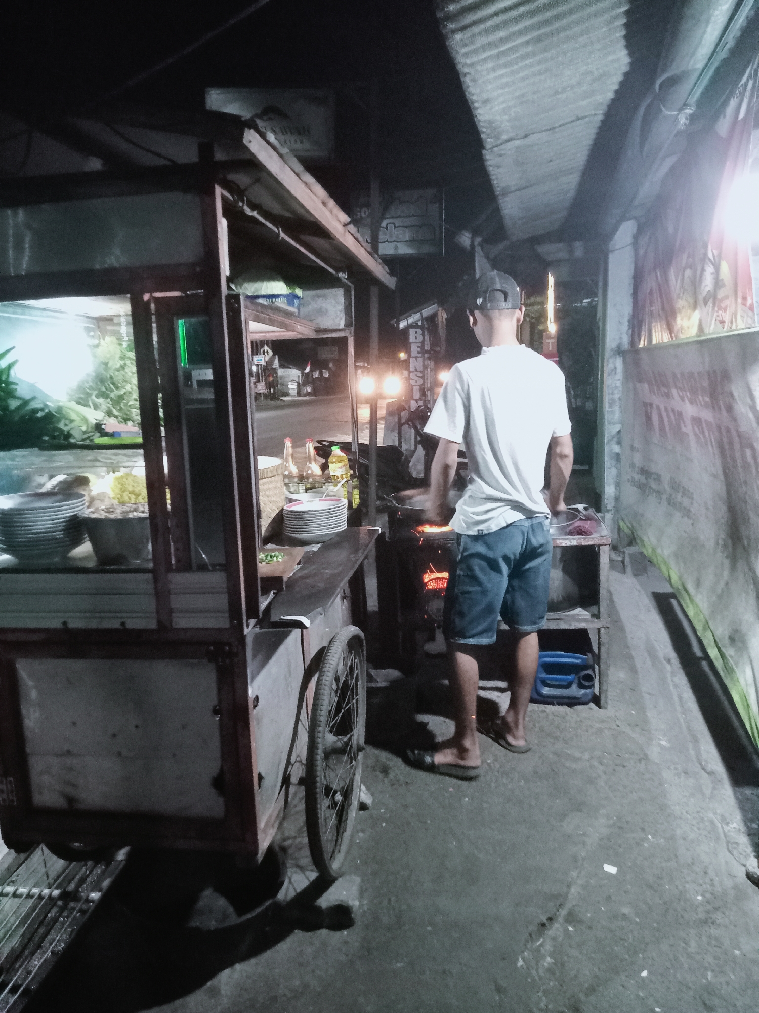In Prajeksari, Indonesia, at night, a man wearing a white t-shirt and denim shorts stands in front of a food cart. He faces away from the camera, appearing to be cooking over a portable stove that emits a visible glow. The food cart, a wheeled structure with a glass display and shelves, is stocked with bowls and stacks of plates. Various bottles, possibly condiments, are also visible on the cart. The scene is illuminated by overhead lights and the glow from the stove, casting shadows on the ground. The environment suggests a street-side vendor operation, with a corrugated metal roof structure extending overhead and a banner on the right side of the frame with Indonesian text. A blue plastic container is visible on the ground near the man's feet.