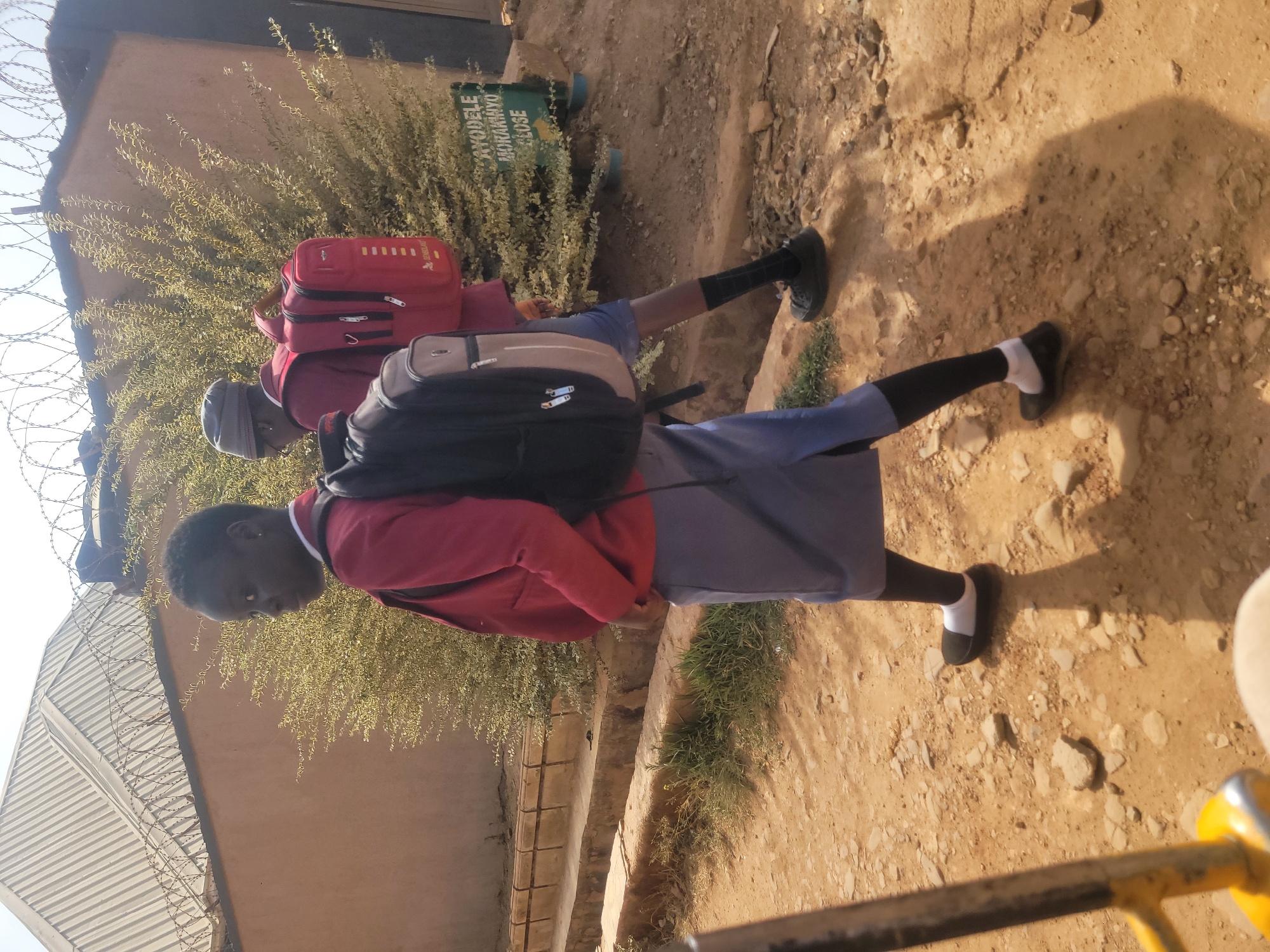 Two young individuals, likely students, are depicted walking outdoors on a rough, unpaved path made of dirt and small stones. Both are wearing school uniforms, which include red long-sleeved tops and blue shorts or skirts. They are carrying backpacks; the person in the foreground has a black and grey backpack, while the one behind carries a red backpack. One student wears black shoes with black socks, while the other wears black shoes with white socks.
The setting features a light brown wall or building behind a line of green, leafy bushes. A barbed wire fence runs along the top of this boundary, suggesting security. In the upper left background, another building with a corrugated metal roof is visible, also topped with barbed wire. The bright sunlight casts distinct shadows on the ground, indicating a clear, sunny day. A yellow metal bar or railing is partially visible in the lower right foreground.
Amidst the bushes, a green sign is visible with white text. The text on the sign reads: "AYODELE", "AKORANMIYO", and "CLOSE". The overall scene portrays a typical outdoor environment in a location like Jos, Nigeria, possibly depicting students on their way to or from school.