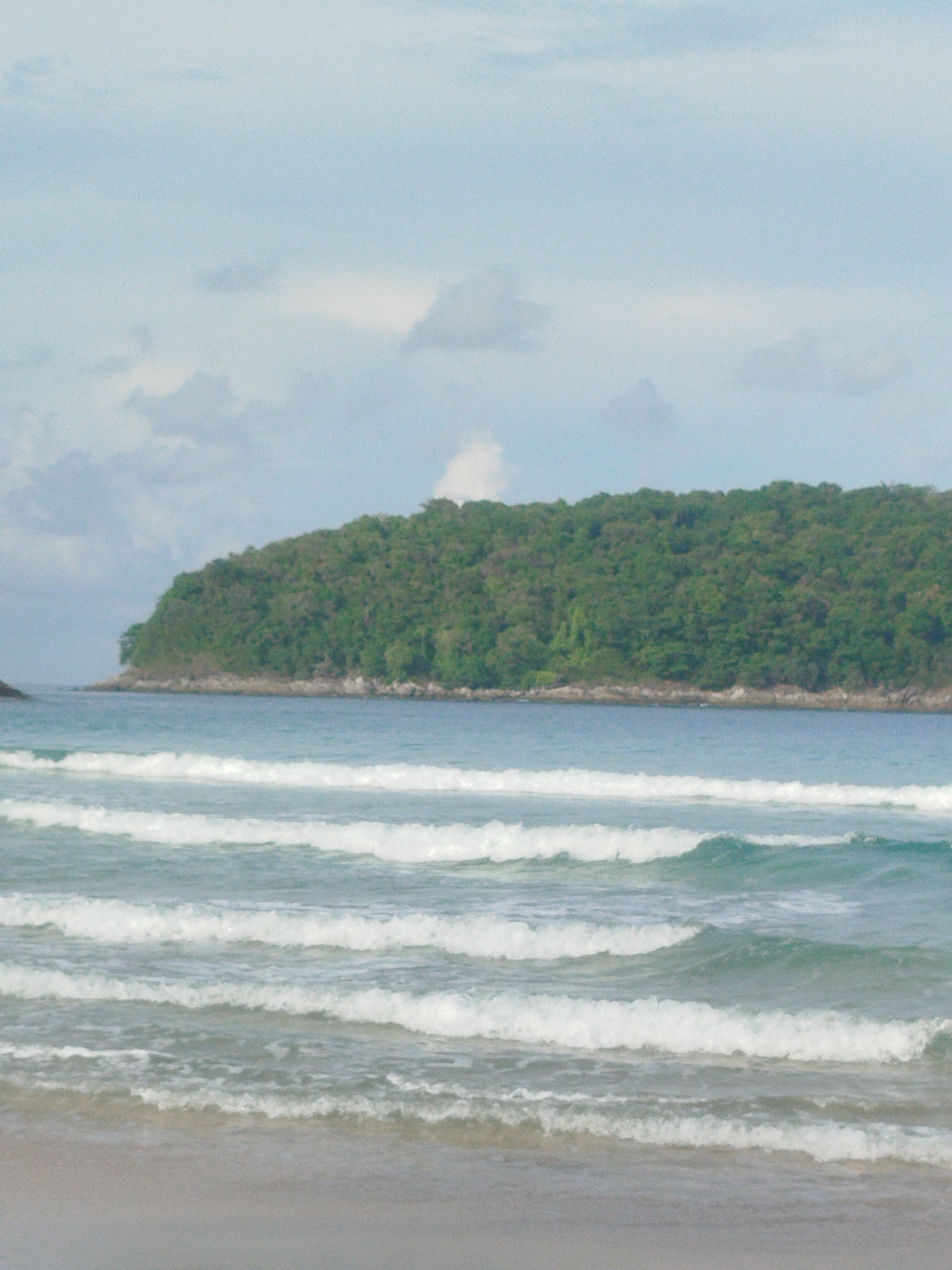 The image depicts a serene beach scene in Rawai, Thailand. The foreground shows a sandy beach leading to the gentle waves of the ocean. Multiple lines of white-capped waves are rolling towards the shore, suggesting a calm but active sea. In the middle ground, the clear blue water stretches towards a lush, green, tree-covered island or peninsula that juts out into the sea. The island appears to have a rocky base along its coastline. The sky above is a pale blue, with scattered, soft clouds, indicating fair weather. No people or man-made structures are visible in the image, emphasizing the natural beauty of the location. The overall impression is one of tranquility and natural coastal scenery.