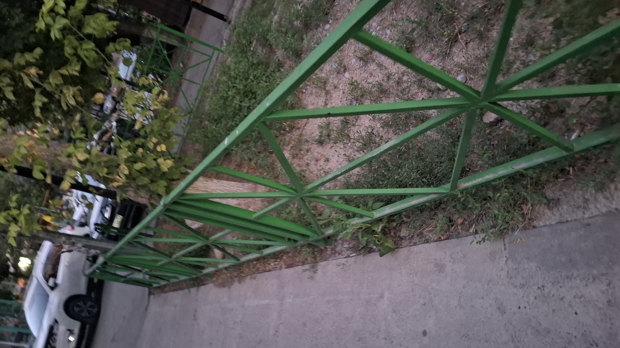 The image shows a green metal fence in a park or along a sidewalk in Shymkent, Kazakhstan. The fence is made of intersecting diagonal bars forming a geometric pattern. To the left of the fence is a concrete sidewalk. Behind the fence, there is dry ground with sparse vegetation and trees with green and yellowing leaves. In the background, several cars are parked or driving, including a white SUV and a dark sedan. The overall lighting suggests it is either early morning or late evening.