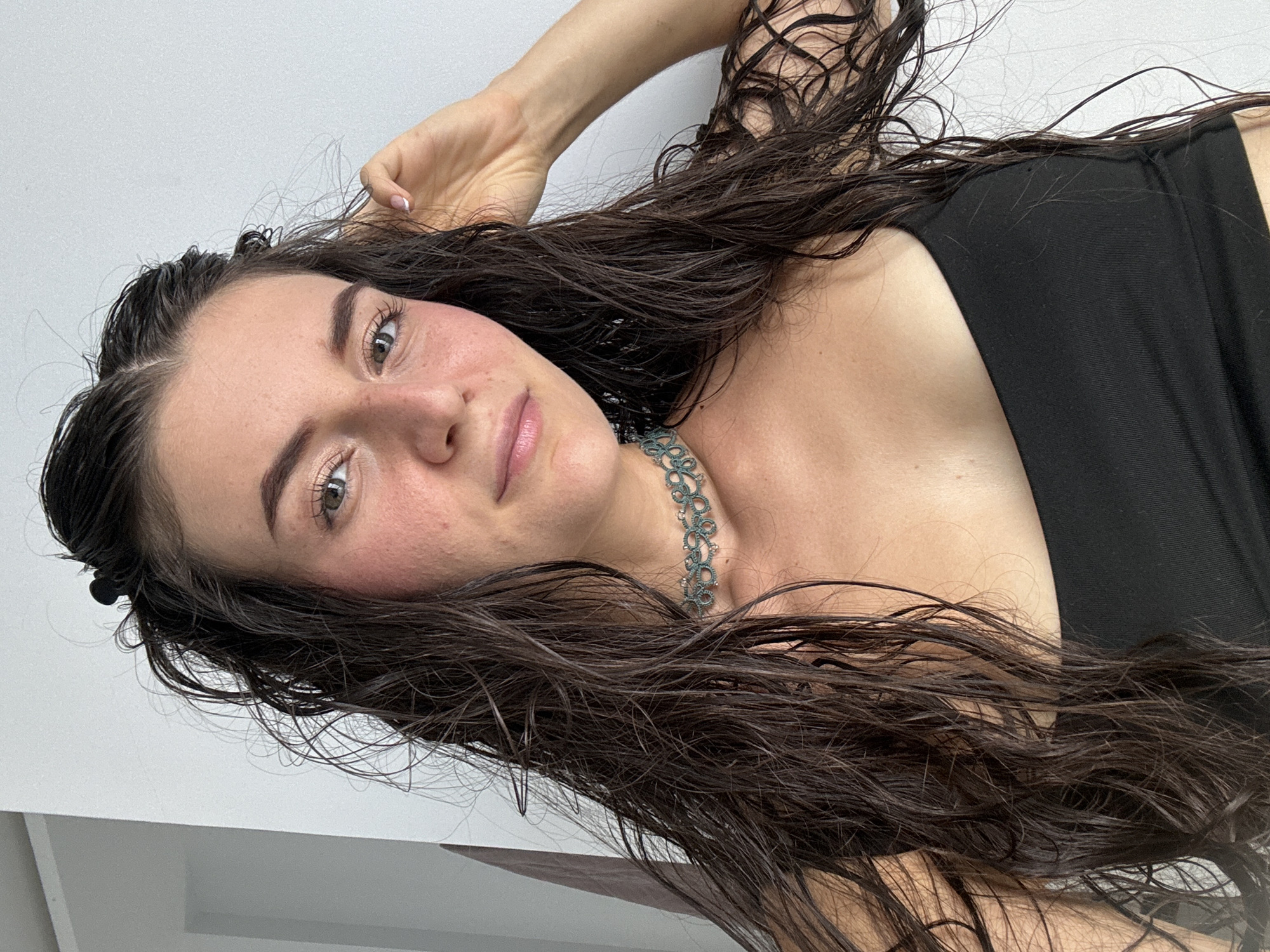 A young woman with long, wet, dark brown hair is facing the camera. She has light skin, pink cheeks, brown eyes, and lightly tinted eyebrows. She is wearing a black tank top and a delicate, greenish-blue choker necklace. Her left hand is raised near her head, fingers touching her hair. The background is a plain, off-white wall and floor. The location is Mũi Né, Vietnam, although there are no specific location markers visible in the image itself.