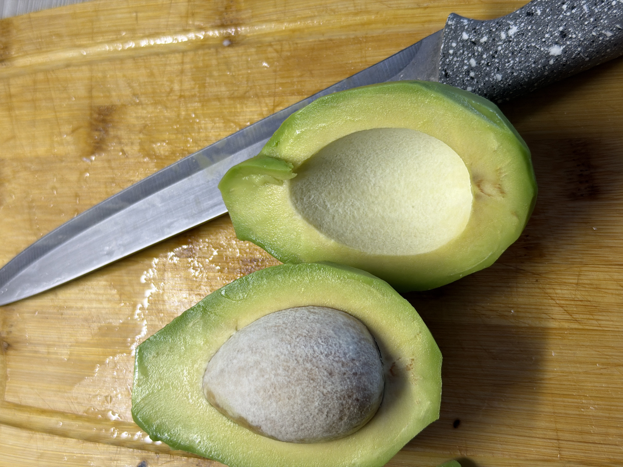 An overhead shot shows a wooden cutting board with a halved avocado and a knife. The avocado is positioned to the left, with the seed still in one half and the other half hollowed out. The knife, with a metallic blade and a speckled gray handle, lies to the right of the avocado. The cutting board exhibits a textured surface with visible wood grain and water droplets. There are no people or any indication of the city of Chaikovsky, Russia, in the image.