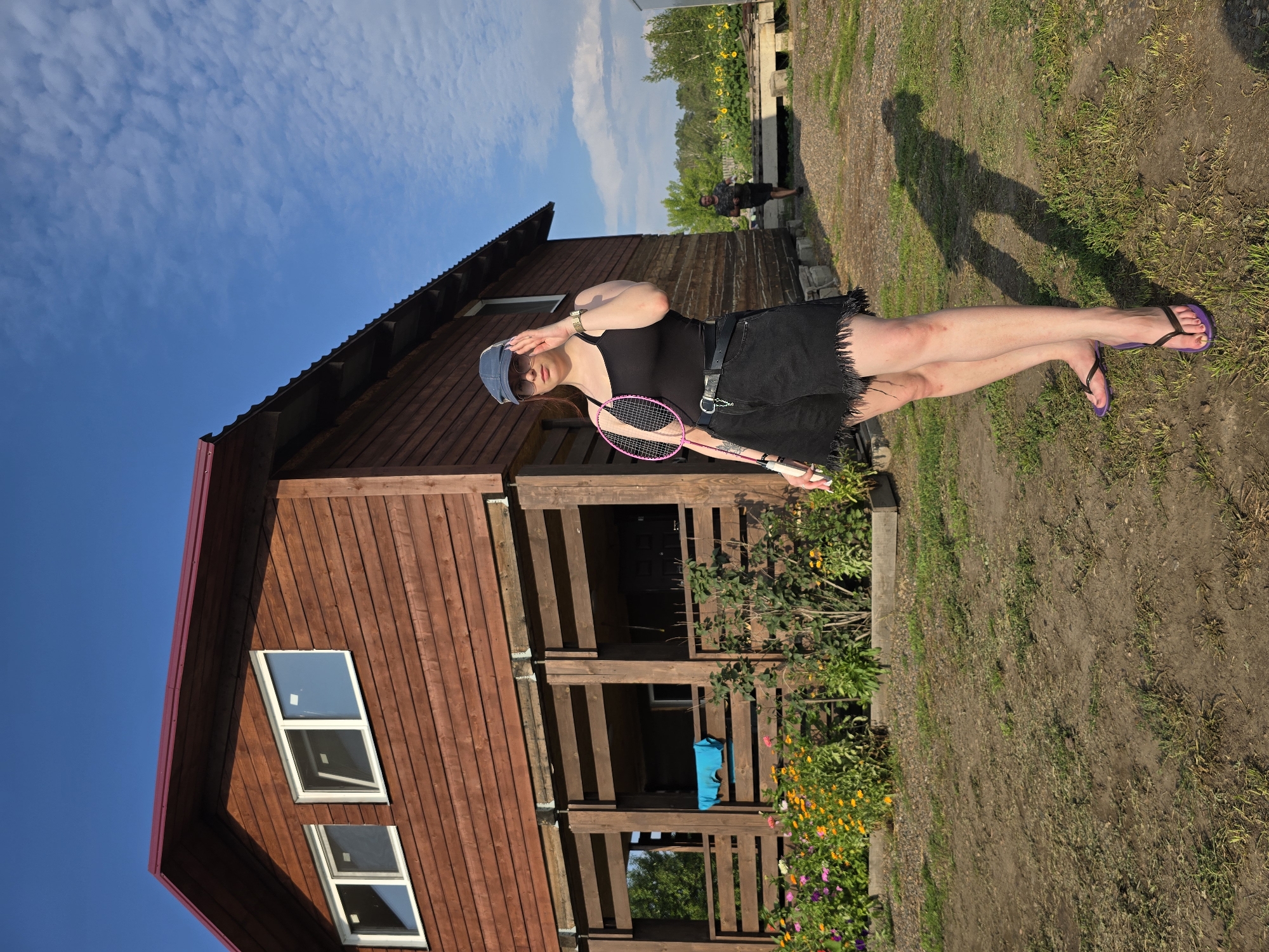 In the village of Yermolayevo, Russia, a young woman stands in front of a two-story wooden house under a bright blue sky with scattered clouds. She is wearing a black tank top, distressed black denim shorts with a frayed hem, a blue baseball cap, and purple flip-flops. She is holding a pink and white badminton racket. Her right hand is raised to adjust her sunglasses. A man in a dark shirt and shorts is visible in the background, walking away from the camera towards a field of sunflowers. The house has a red metal roof and wooden siding. A porch with wooden railings is visible on the first floor, with a blue towel hanging on the railing. A flower bed with various colorful flowers is planted in front of the porch. The ground is a mixture of dirt and sparse grass. The overall scene suggests a casual outdoor setting on a sunny day.