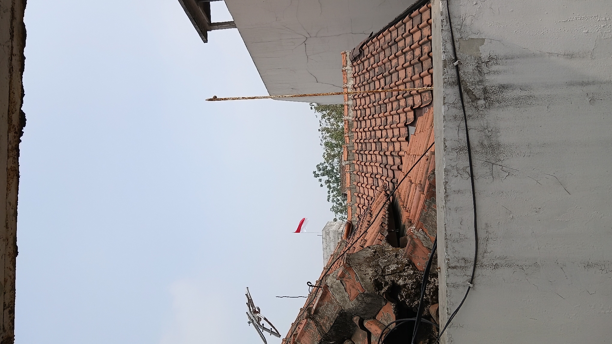 The image shows a partially collapsed tiled roof with exposed brick and debris in what appears to be an urban setting, possibly in Bandung City, Indonesia. A small Indonesian flag is visible flying from a rooftop in the background. The scene is viewed from behind a weathered concrete wall with peeling paint, from which black cables are strung. The sky is clear and pale blue, and trees are visible behind the damaged roof. The overall impression is one of disrepair and possibly storm damage. There are no people visible in the image.