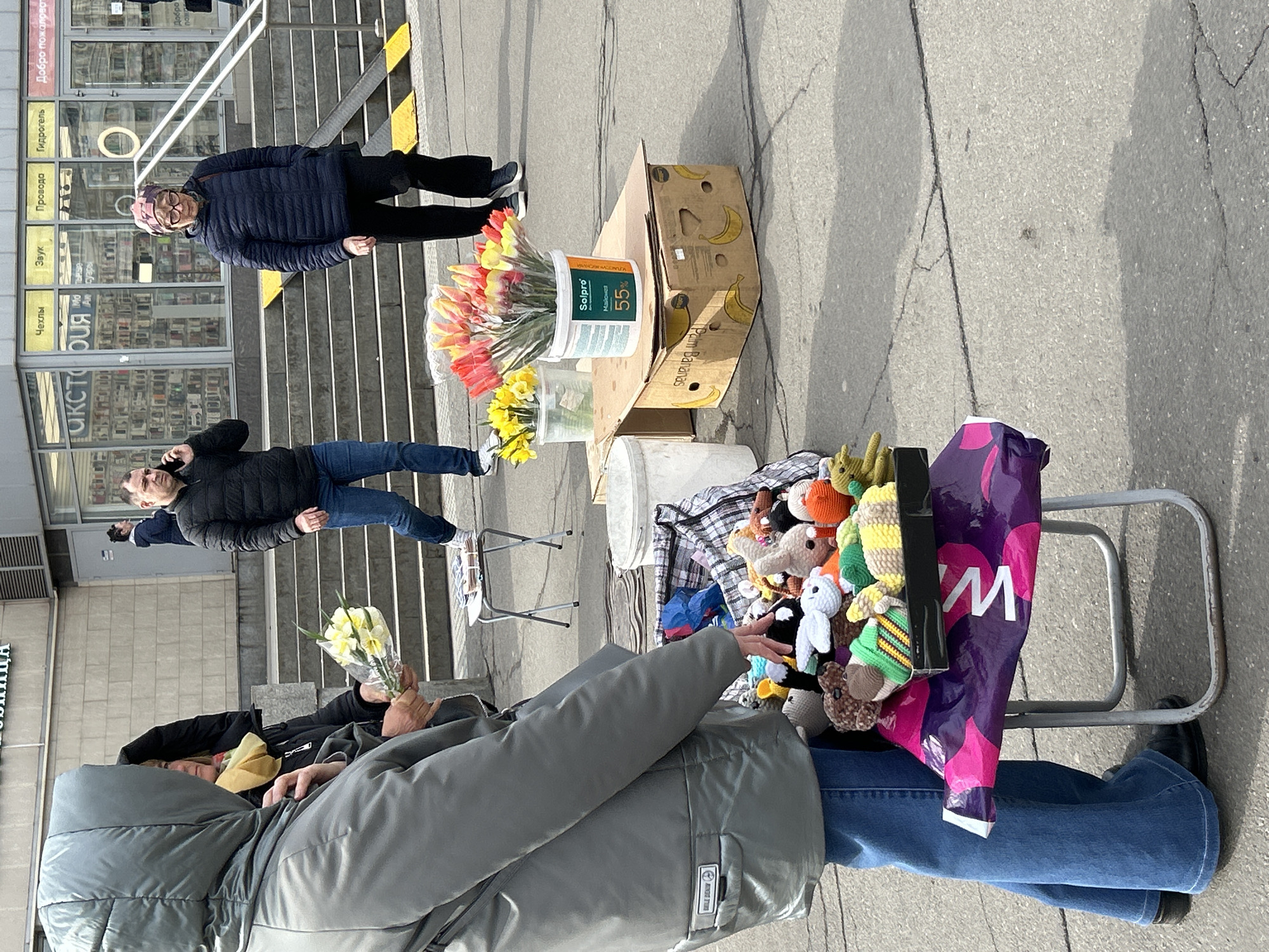 This media file captures an outdoor street scene, likely in Saint Petersburg, Russia, given the visible Cyrillic text. The image is taken during the daytime, with bright sunlight casting clear shadows.
The foreground features a person wearing a grey puffer jacket and blue jeans, seated on a low chair. They are holding a small bouquet of yellow daffodils and reaching towards a display of various colorful crocheted toys, which are arranged on a trolley or stand. A purple shopping bag with a white "W" logo is visible next to the toy display.
To the left of the toy stall, a street vendor setup includes two buckets of flowers: one white bucket filled with red and yellow tulips, and a clear container holding more yellow daffodils. These flowers rest on a repurposed cardboard box labeled "Palm Bananas." Adjacent to this, another white bucket bears a label in Cyrillic, reading "Solpro Майонез 55%" (Solpro Mayonnaise 55%).
In the middle ground, a concrete staircase leads up to a building entrance. A man in a dark puffer jacket and blue jeans stands on the stairs, talking on a mobile phone. Further up the stairs, an older woman wearing a dark blue puffer jacket and a patterned head covering stands and looks towards the camera.
The background shows the glass facade of a building, reflecting its surroundings. Several signs with Cyrillic text are visible on the building, including "Добро пожаловат" (Welcome), along with terms like "Гидрогель" (Hydrogel), "Провода" (Wires), "Звук" (Sound), and "Чехлы" (Cases), suggesting an electronics or accessories store. The overall scene depicts a bustling urban area with street vendors and passersby.