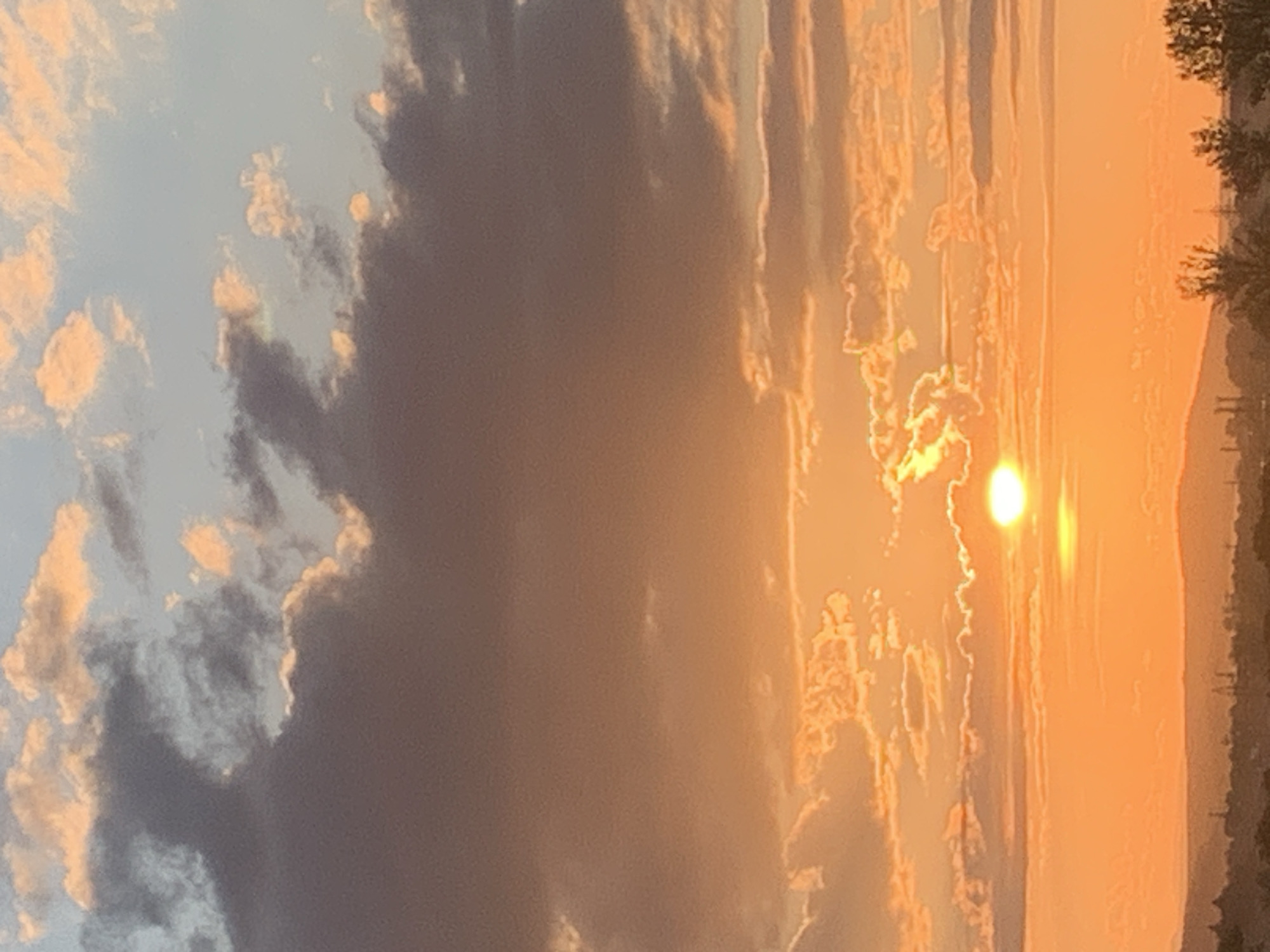 This image captures a sunset over the city of Kolomyia, Ukraine. The sky is a vibrant display of orange and yellow hues, with scattered clouds ranging from light pink to dark gray. The sun, a bright yellow orb, is partially obscured by a thin cloud layer near the horizon. Below the sky, a silhouetted landscape of hills, trees, and industrial structures forms the city's outline. The scene suggests a peaceful end to the day.