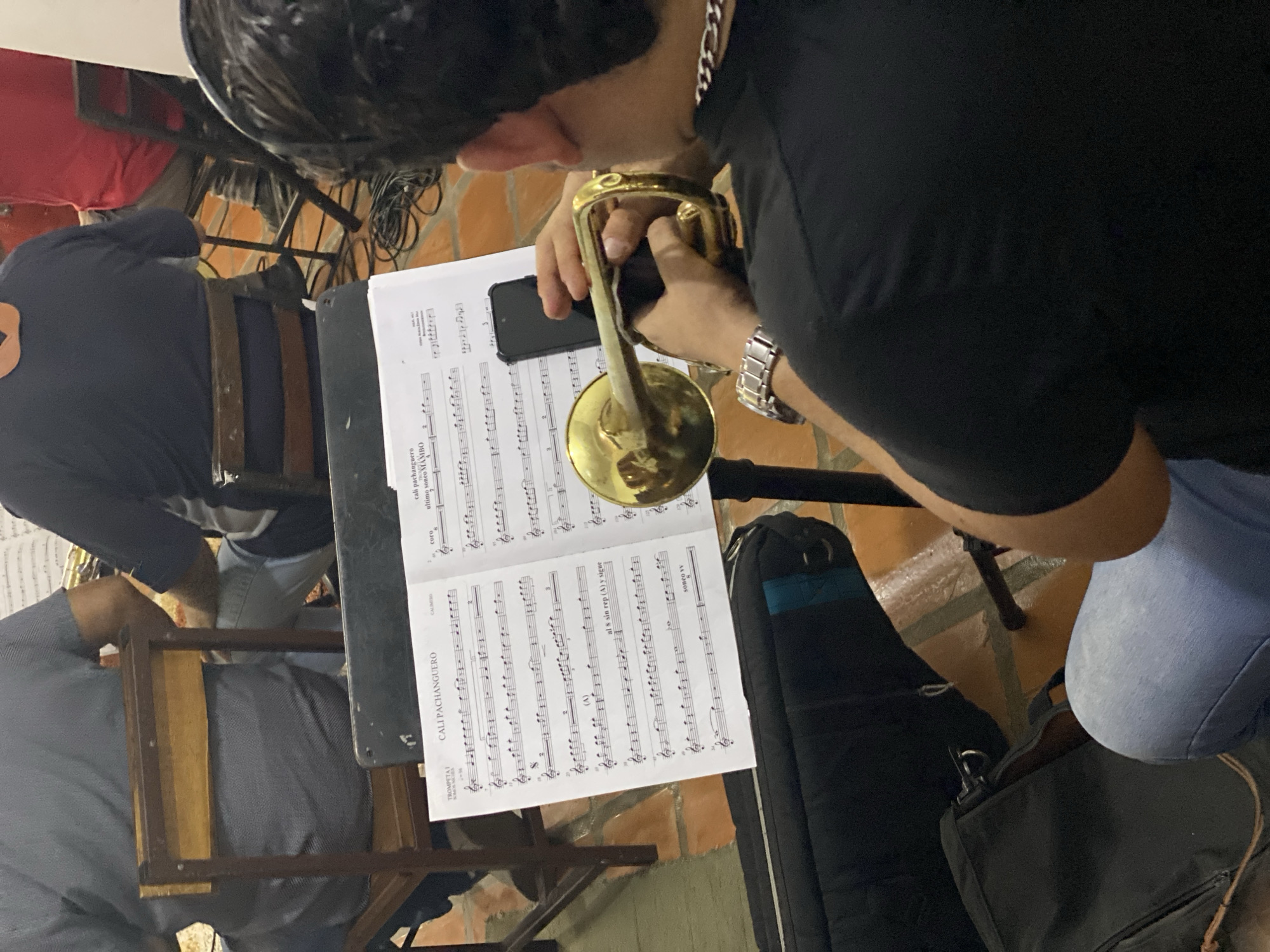 A group of musicians is observed indoors, likely during a rehearsal or preparation for a performance in Tinaquillo, Venezuela. The image is taken from an overhead perspective.
The central figure, a man with dark hair, wearing a black t-shirt, a silver chain, and a silver watch, is focused on a golden brass instrument, likely a trumpet, which he holds with both hands. His posture suggests he is meticulously tuning, cleaning, or getting ready to play the instrument.
Directly in front of him is a music stand holding an open sheet of music. A black smartphone is resting on the sheet music. The sheet music contains musical notation and several visible text elements, including "TROMPETAI" (indicating Trumpet 1 part), "CALI PACHANGUERO" (the likely title of the piece, appearing multiple times), "coro," "ultimo sonco MAMBO," "(A)," "al & sin rep (A) y sigue," and "soneo vv." These phrases suggest a Latin American music piece, possibly a Mambo, with specific instructions for the musician.
Two other individuals are partially visible, seated on wooden chairs. One, to the left of the main subject, is seen from the back, wearing a dark blue long-sleeved shirt and blue jeans. Another person's arm is slightly visible further left.
The floor is covered with reddish-brown tiles. A black instrument case or bag rests on the floor to the right of the music stand. Various cables are visible on the floor and in the background, indicating the presence of sound or electrical equipment. The setting appears to be a casual indoor space, possibly a practice room or a small venue, with a focus on the musicians' readiness to play.