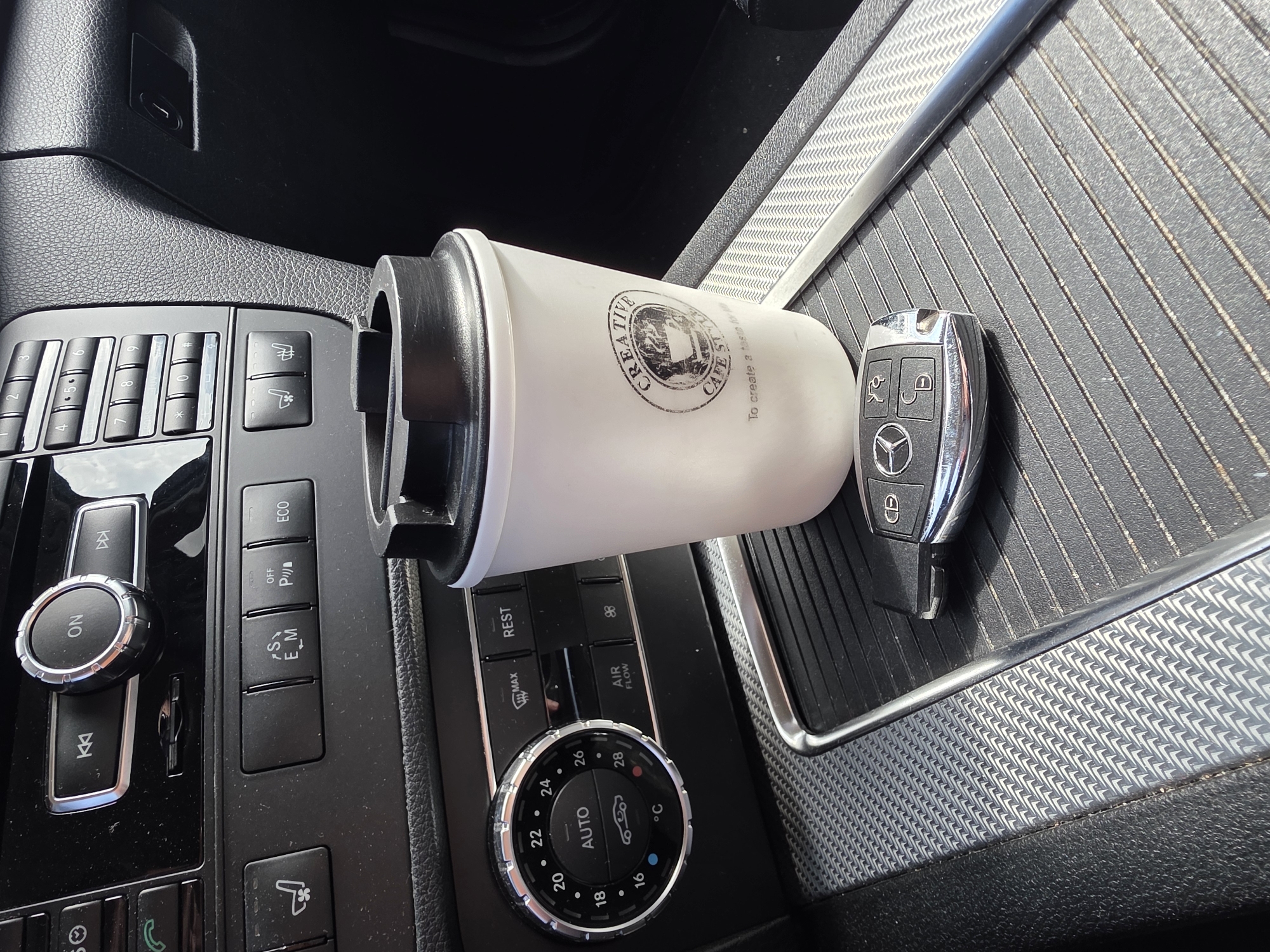 The image depicts the interior of a car, specifically the center console area. A white coffee cup from "Creative Cafe Style" is positioned horizontally between the car's control panel and a compartment with a textured surface. A car key with the Mercedes-Benz logo lies next to the coffee cup. The control panel features various buttons and dials, including climate control settings (temperature ranging from 16°C to 28°C and an "AUTO" setting) and buttons labeled "ECO," "S E_M," and "OFF P" with a parking symbol. The location in Plovdiv, Bulgaria is not identifiable from the details in the image.
