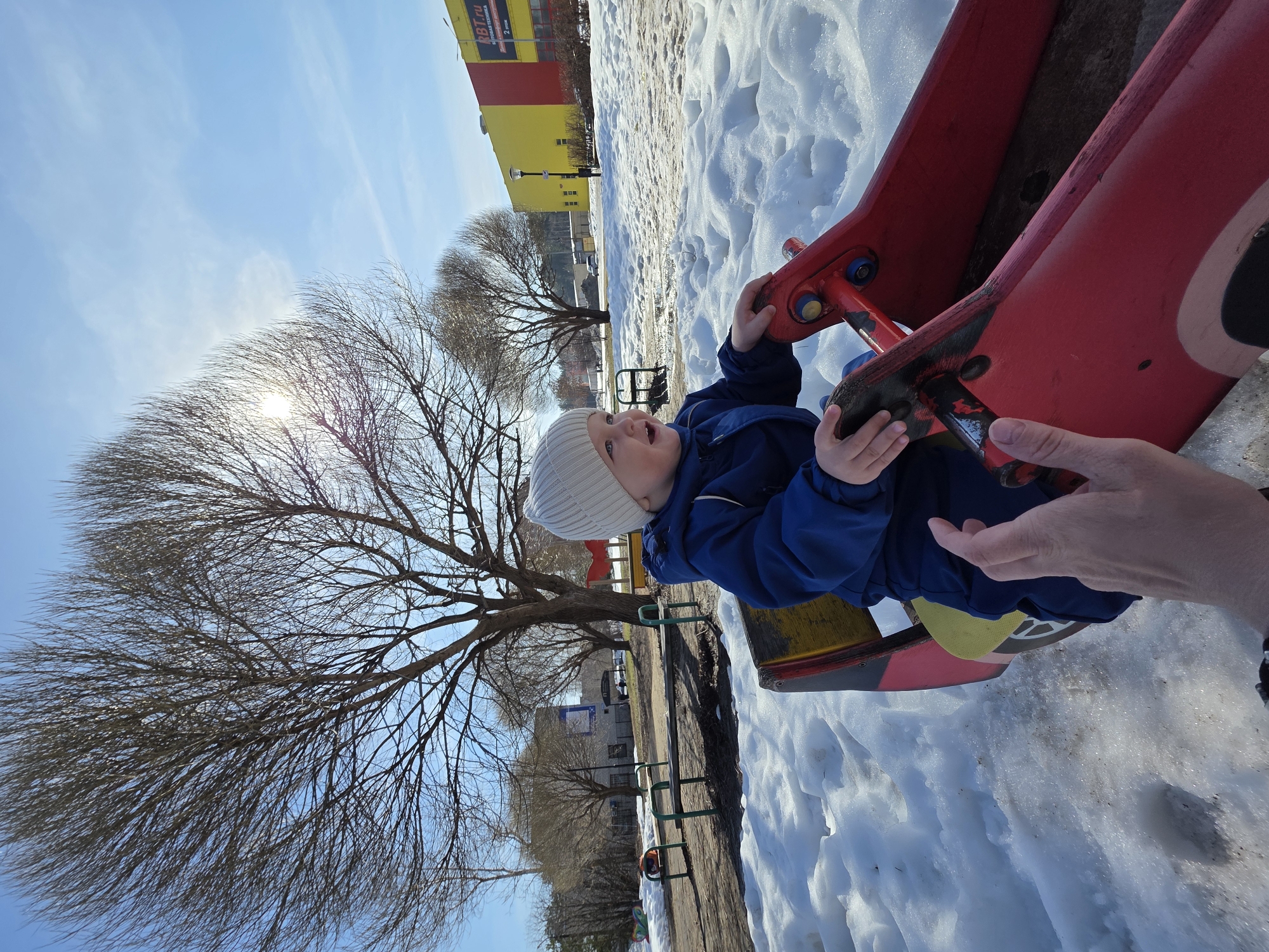 On a bright day in Chaikovsky, Russia, a young child wearing a blue snowsuit and a white knit hat is sitting on a red and yellow seesaw in a snow-covered park. The child is holding onto the handles of the seesaw and appears to be smiling or laughing. A person's hand is visible, seemingly assisting or supervising the child. In the background, there is a building with a red and yellow facade, bare trees, and patches of snow on the ground. Other playground equipment can be seen in the distance. The sun is shining brightly, partially obscured by the branches of a large tree.