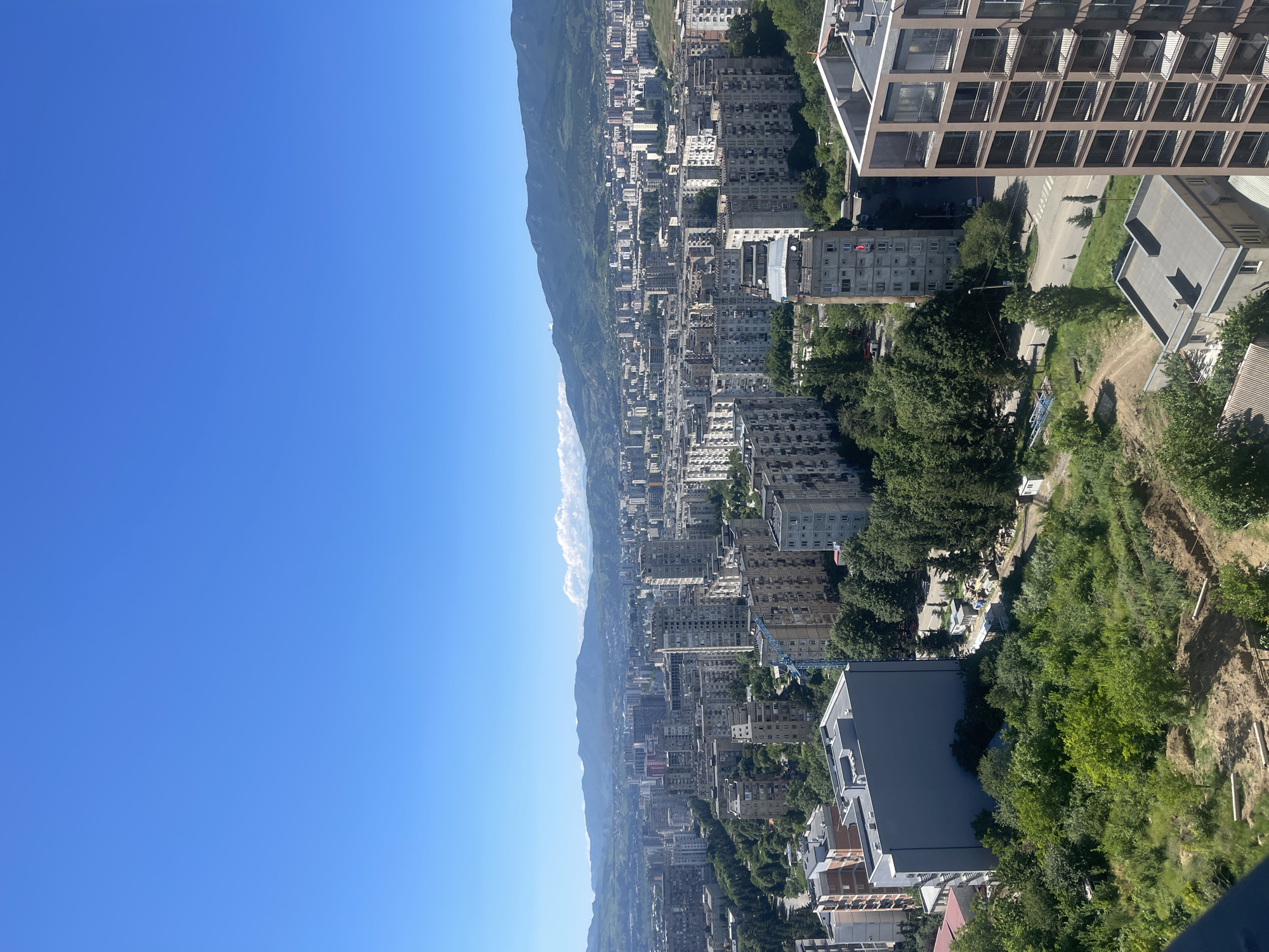 An aerial wide shot captures a sprawling urban landscape under a clear blue sky in Tbilisi, Georgia. The city is densely packed with apartment buildings of varying heights and architectural styles, many appearing to be of Soviet-era construction with their utilitarian grey facades. In the foreground, a modern apartment building with a glass and metal exterior is partially visible on the right. To the left, a large, plain grey building stands out. Green trees are interspersed throughout the city, and a construction site with earthworks and piping is visible in the lower left quadrant. A blue construction crane is positioned among the buildings. In the distance, rolling hills and mountains form a natural backdrop to the city, with a line of white clouds near the mountain peaks. The scene depicts a sunny day in a developed urban area.