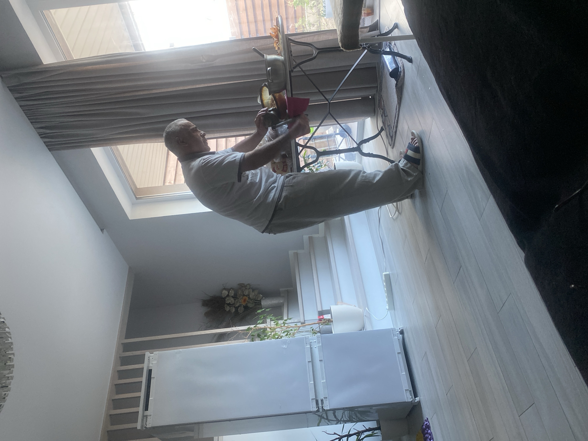 A man in a grey t-shirt and light grey pants is performing a handstand in what appears to be a modern living room in Novi Petrivtsi, Ukraine. He is upside down, with his feet in the air and his hands on the floor. He is holding a red book in his right hand. The room has a high ceiling with a skylight, and the floor is covered with light-colored wooden planks. There are stairs in the background, and a large white appliance, possibly a refrigerator, is visible on the lower level. A decorative bouquet of flowers is placed on a shelf near the stairs. The sunlight streaming through the skylight suggests it is daytime. The man appears focused and in control of his balance.