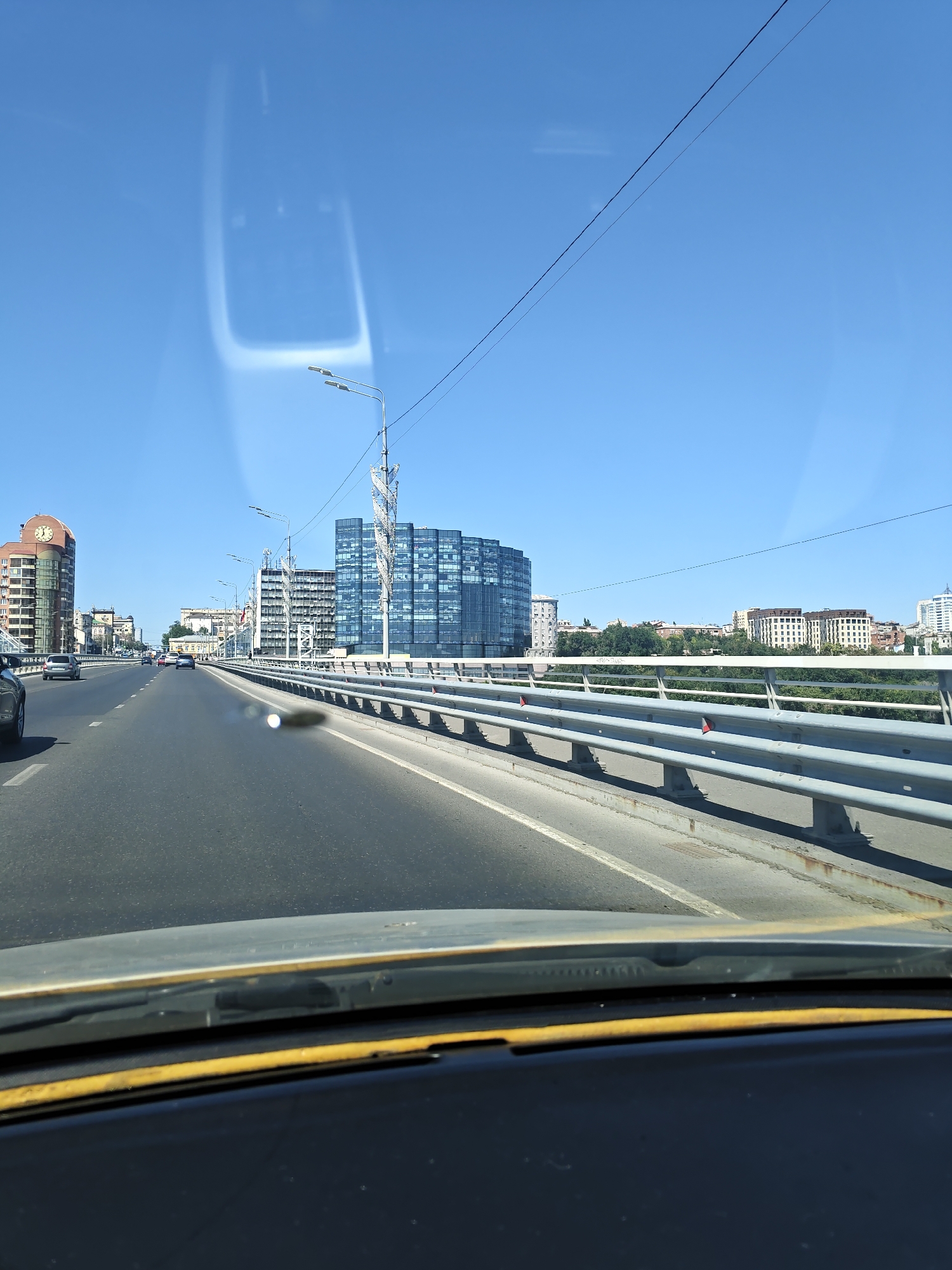 The image captures a perspective from a vehicle traveling on a bridge over a river in Rostov-on-Don, Russia. The sky is clear blue. On the left side of the bridge, there are several buildings, including a tall, modern building with a clock face and other residential buildings. On the right side of the bridge, there are more modern buildings, some with glass facades, and a line of trees and buildings in the distance. The bridge itself has a metal railing along its edge and streetlights. The road appears to be a multi-lane highway. Reflections are visible on the windshield of the vehicle. The scene depicts a typical urban cityscape during the day.