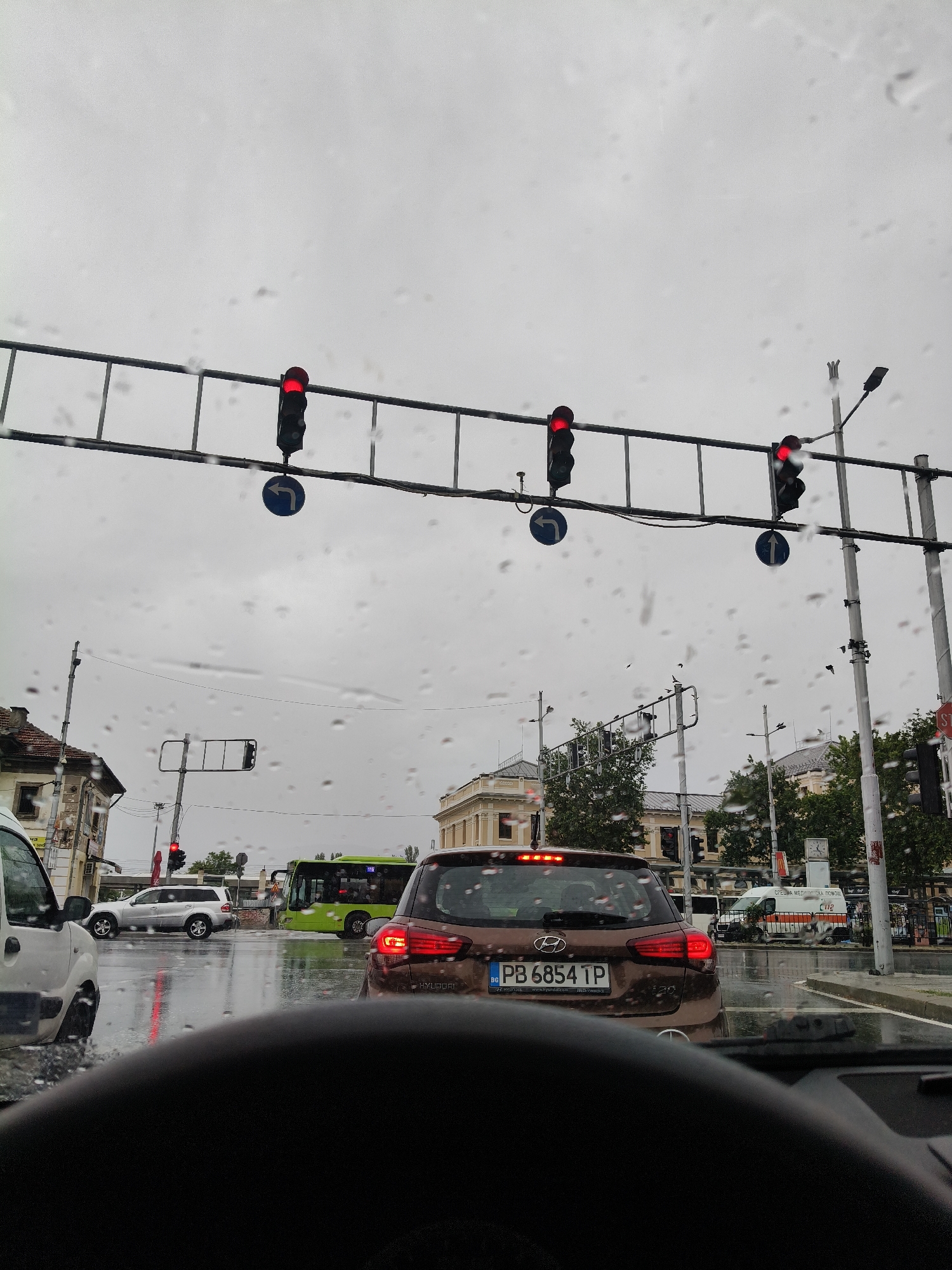 The image depicts a street scene in Plovdiv, Bulgaria, on a rainy day. The perspective is from inside a vehicle, looking forward through a rain-streaked windshield. Multiple traffic lights are visible, all showing red, indicating a stop. In the foreground, the dashboard of the car is visible, along with the back of a brown Hyundai hatchback. A white van is parked to the right, and a green bus is in the middle distance, also stopped. Several other vehicles, including a silver SUV, are also present and stationary. Buildings line the street, some with visible architectural details. The sky is overcast and gray, consistent with rainy weather. The street surface is wet and reflects the surrounding lights. The overall impression is of traffic congestion at an intersection during a downpour.