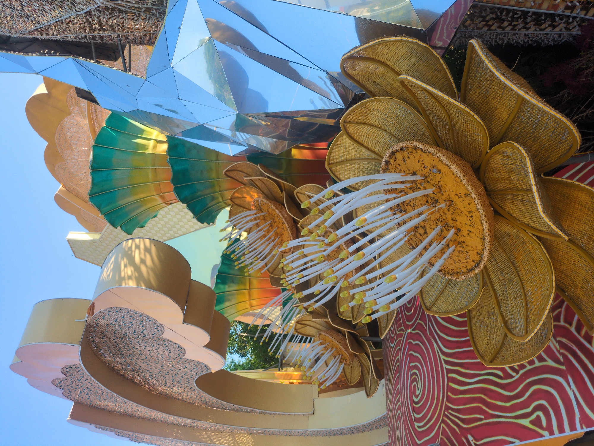 An eye-level, close-up shot depicts a large, flower-themed art installation with reflective panels, set against a clear blue sky. The installation features oversized, golden yellow flowers with textured petals and prominent white stamens. Some petals have a green-to-yellow gradient, and the floral structures are arranged in a layered fashion. Mirrored panels with sharp angles reflect the sky and surrounding environment. The artwork also includes decorative, rounded sections with intricate patterns in neutral tones. The backdrop includes a section with red and white swirl patterns. The location is Tuy Hòa, Vietnam, as the installation is part of a public art display in the city.