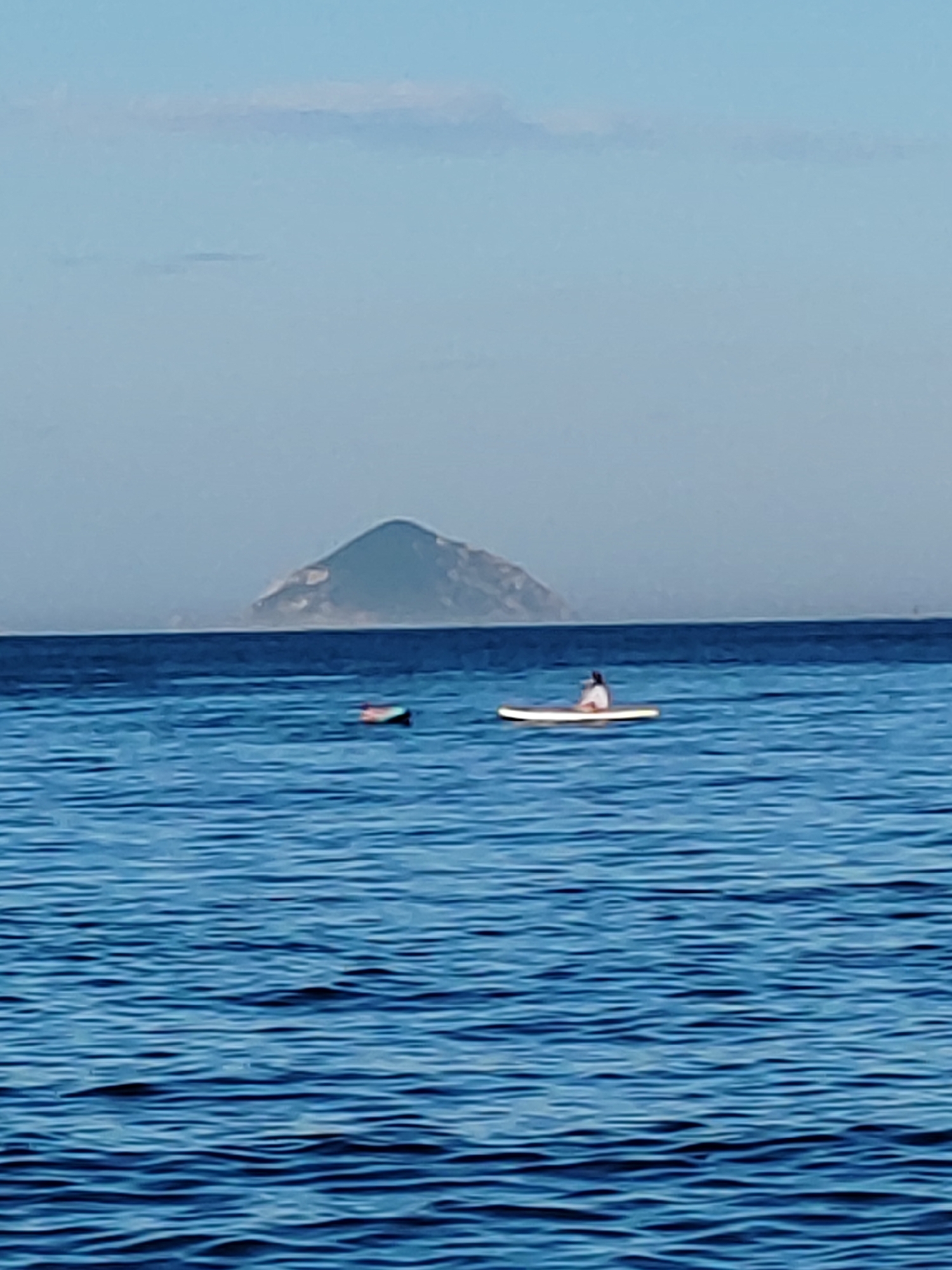 A wide shot captures a tranquil scene on the ocean with a distant, hazy mountain range and a clear blue sky. In the foreground, the deep blue water is disturbed by gentle ripples. Two stand-up paddleboards are visible; one closer to the left is red and white and appears unoccupied. Further to the right, another paddleboard is occupied by a person wearing a white top and dark bottoms, sitting down on the board. The overall impression is one of peaceful recreation in a coastal environment, potentially near Nha Trang, Vietnam, given the characteristic geological formations of the region and the warm, humid atmosphere suggested by the haze.